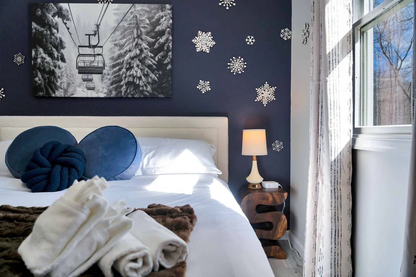 A bedroom decorated with snowflake wall decals, a black and white mountain and ski lift artwork, and a window with white curtains letting in sunlight.