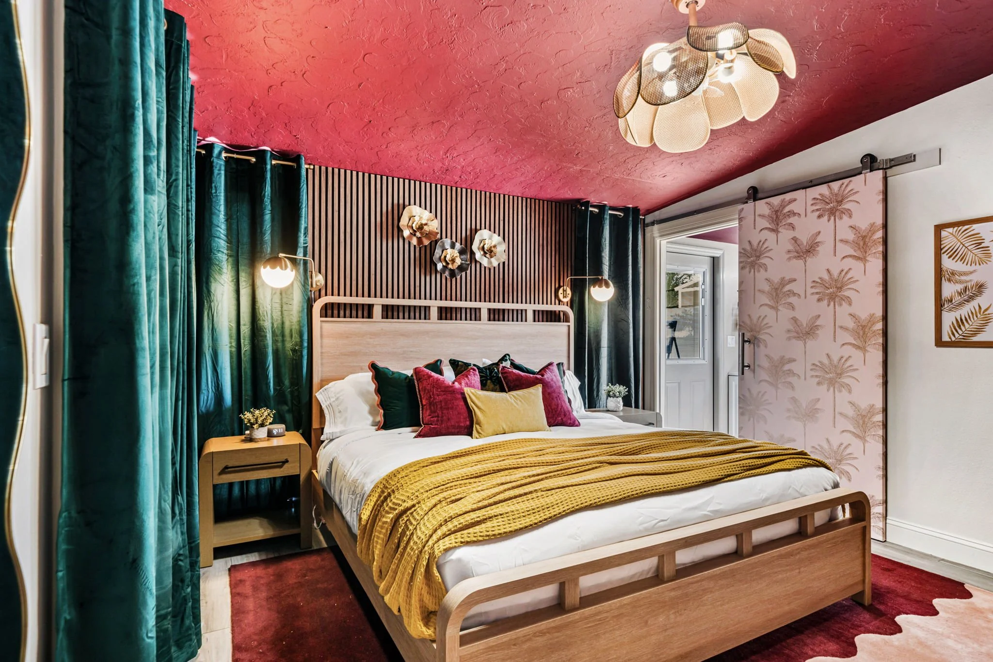 A bedroom with a wooden bed, colorful pillows, a yellow blanket, green curtains, a red ceiling, and floral decor on the wall.