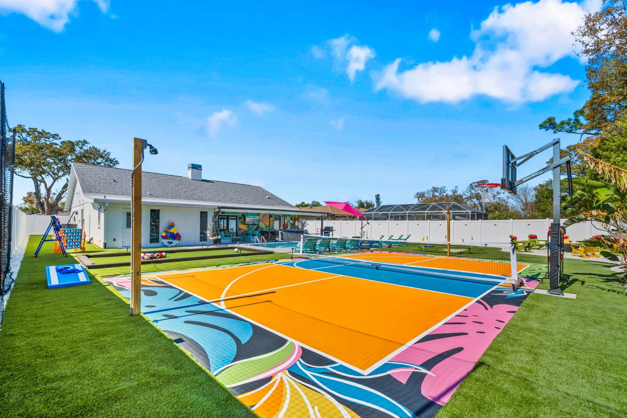 Colorful outdoor sports court with a basketball hoop, surrounded by a backyard with a house, seating area with umbrella, and children's play equipment under a blue sky.