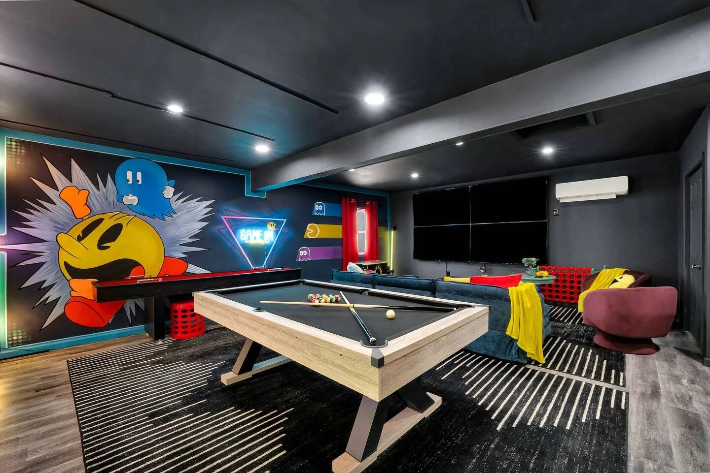A modern entertainment room featuring a Pac-Man mural, a pool table with pool balls and cues, a blue sofa, a pink armchair, a wall-mounted TV, and neon arcade signs.