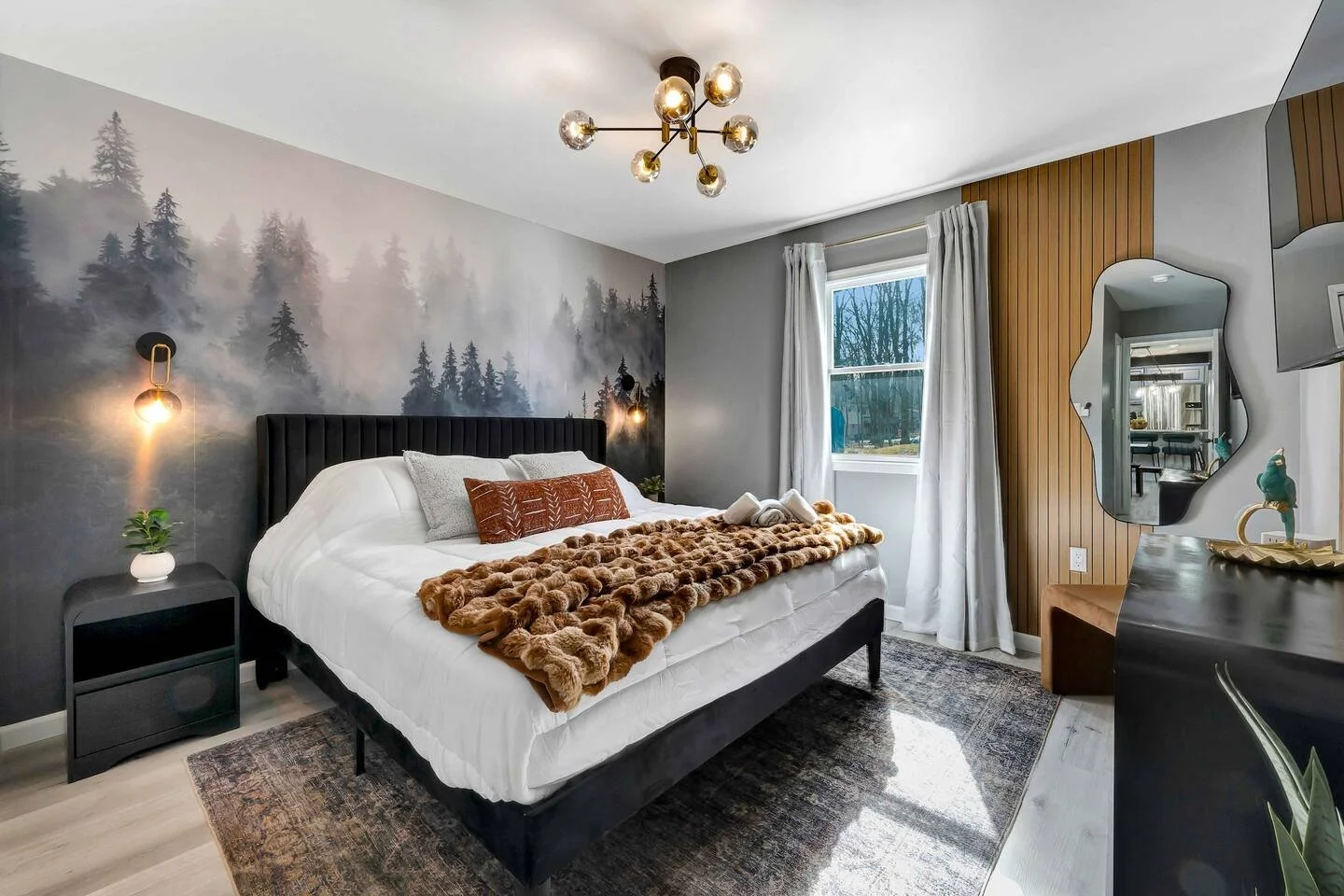 Modern bedroom with a large bed, fuzzy blanket, decorative pillows, wall art of foggy forest, and contemporary lighting fixtures.