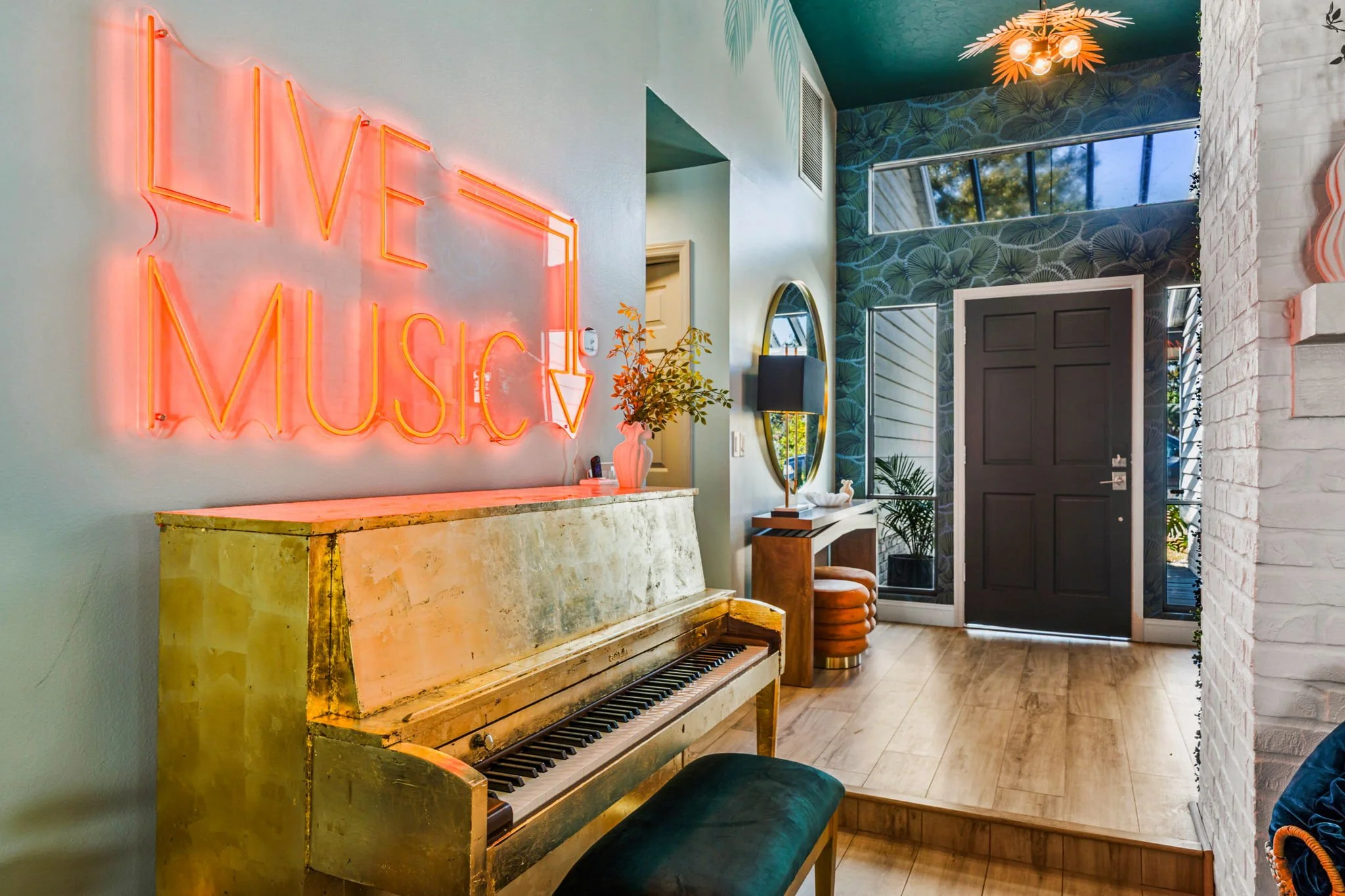Interior of a modern home with a neon sign that says 'Live Music' on the wall, an old gold-colored piano, decorative vases, a mirror, orchids, and a mosaic wall near the entrance door.