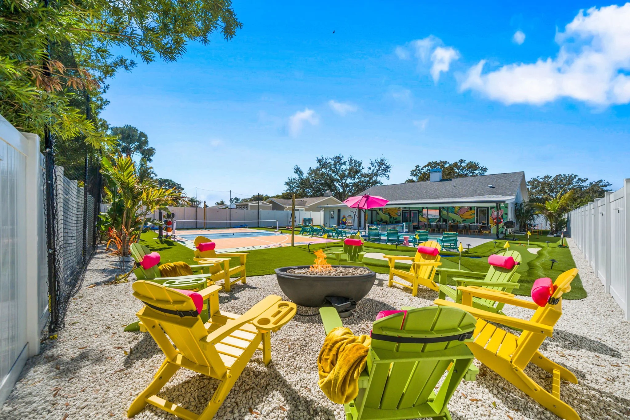 Bright backyard with yellow and green lawn chairs, a fire pit, a swimming pool with a net, and colorful umbrellas under a clear blue sky.