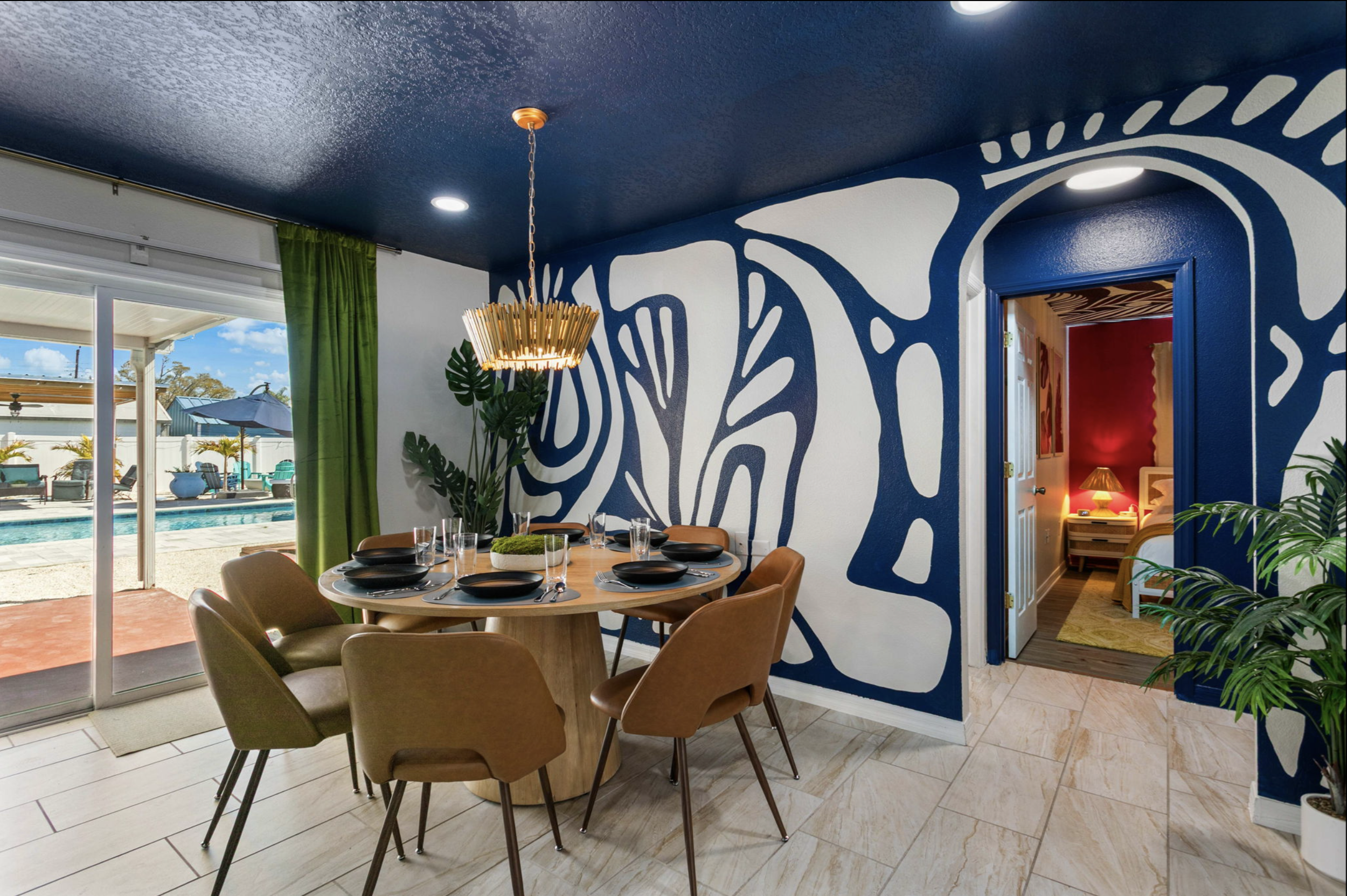 Dining room with a round wooden table set for six, tan chairs, a modern chandelier, and a large wall mural with abstract blue and white patterns. Open door reveals a bedroom with red walls, a nightstand, a lamp, and a bed.