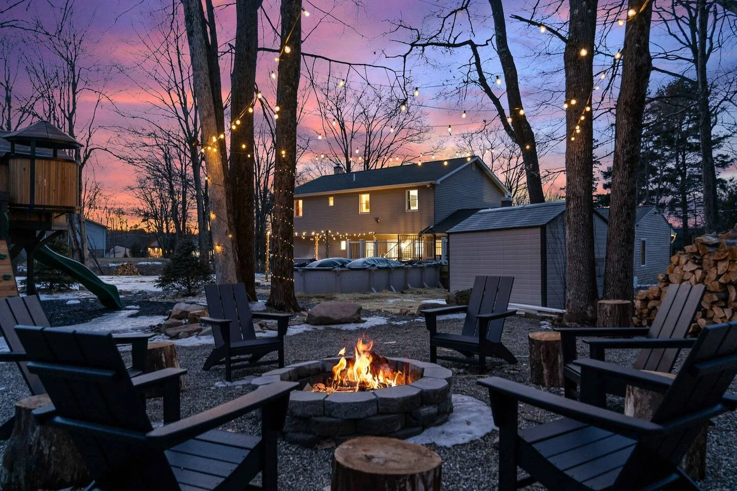 Backyard scene at dusk with a circle of black Adirondack chairs around a stone fire pit, decorated with string lights hanging from trees, and a house with lit windows in the background. There is snow on the ground and a playset with a slide on the le