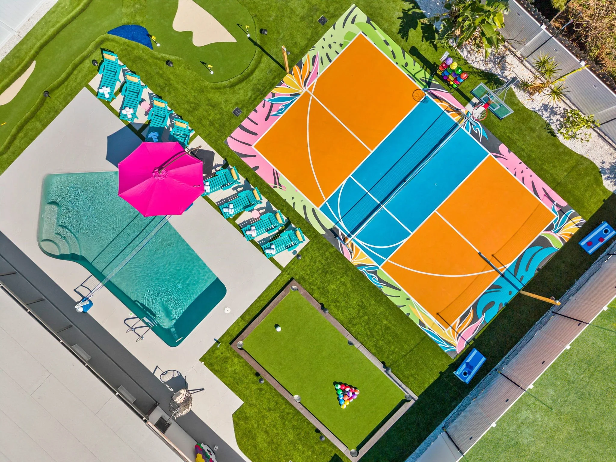 A colorful outdoor recreational area with a swimming pool, a pink umbrella, a section of lounge chairs, a vibrant basketball court, and a small pool table surrounded by green grass and landscaped areas.