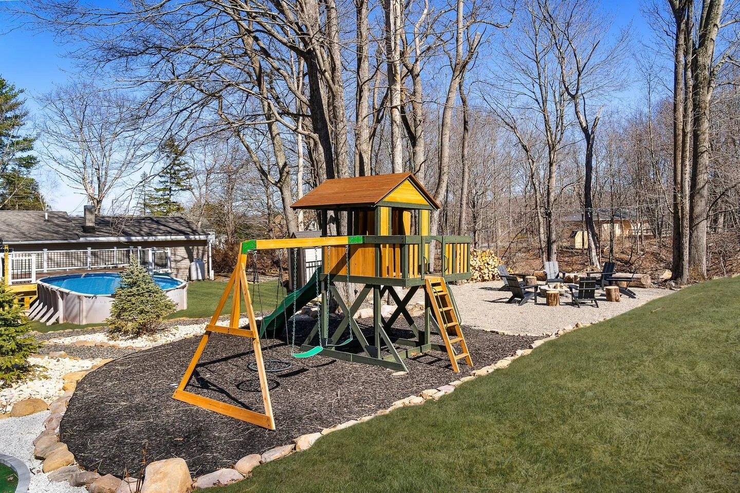 Backyard with a wooden playset, sandbox, above-ground pool, gravel seating area with Adirondack chairs, surrounded by tall trees and a lawn.