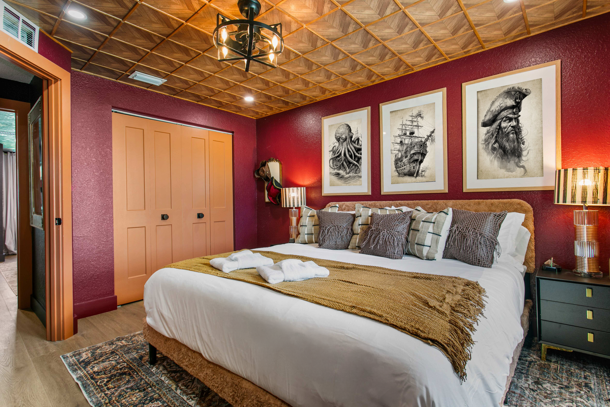 A cozy bedroom with red walls and a wooden coffered ceiling featuring a black chandelier. The bed has white linens, a brown blanket, and an assortment of pillows. On the wall above the bed are three framed black-and-white pirate-themed drawings. Ther