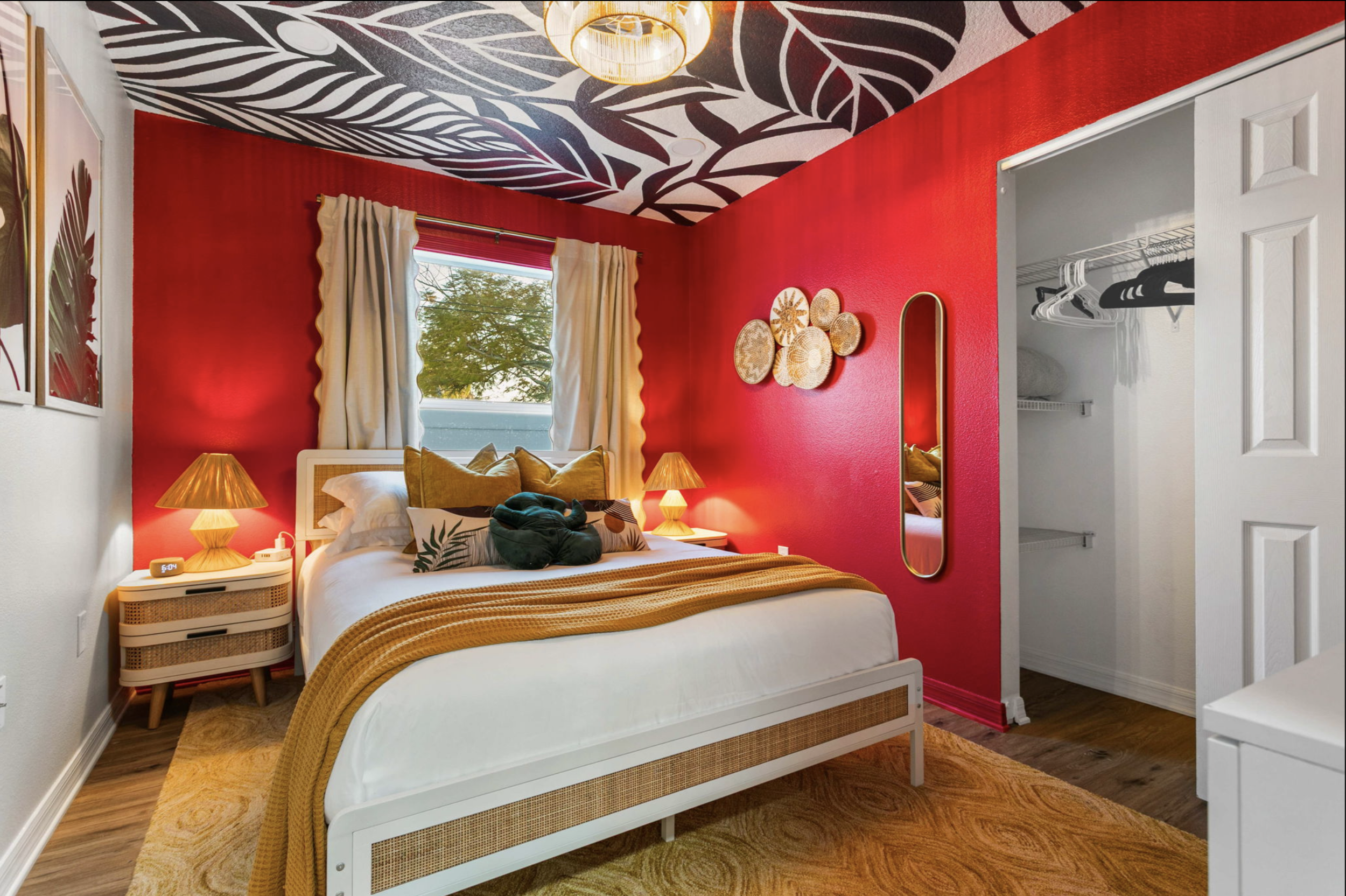 A bedroom with red walls, a window with cream curtains, and a bed with white bedding, multiple pillows, and a green stuffed animal. There are two nightstands with lamps, a decorative wall hanging, a mirror, and an open closet with hangers.