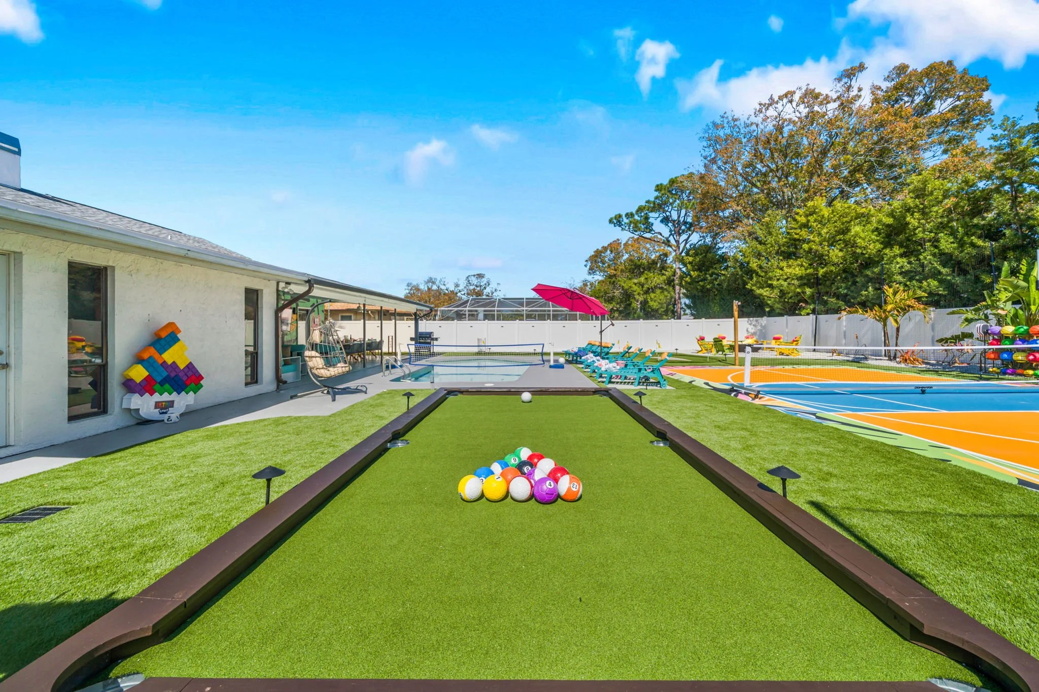 Backyard with pool, outdoor furniture, shady trees, and sports courts under a blue sky.