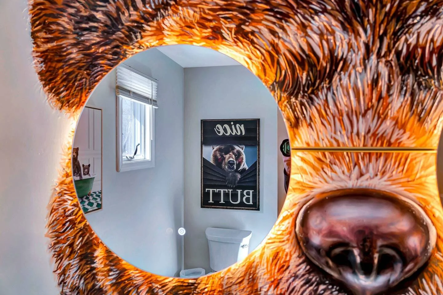 A bathroom mirror with an orange, furry border reflecting a wall with a bear poster, window, and part of a person taking the photo.