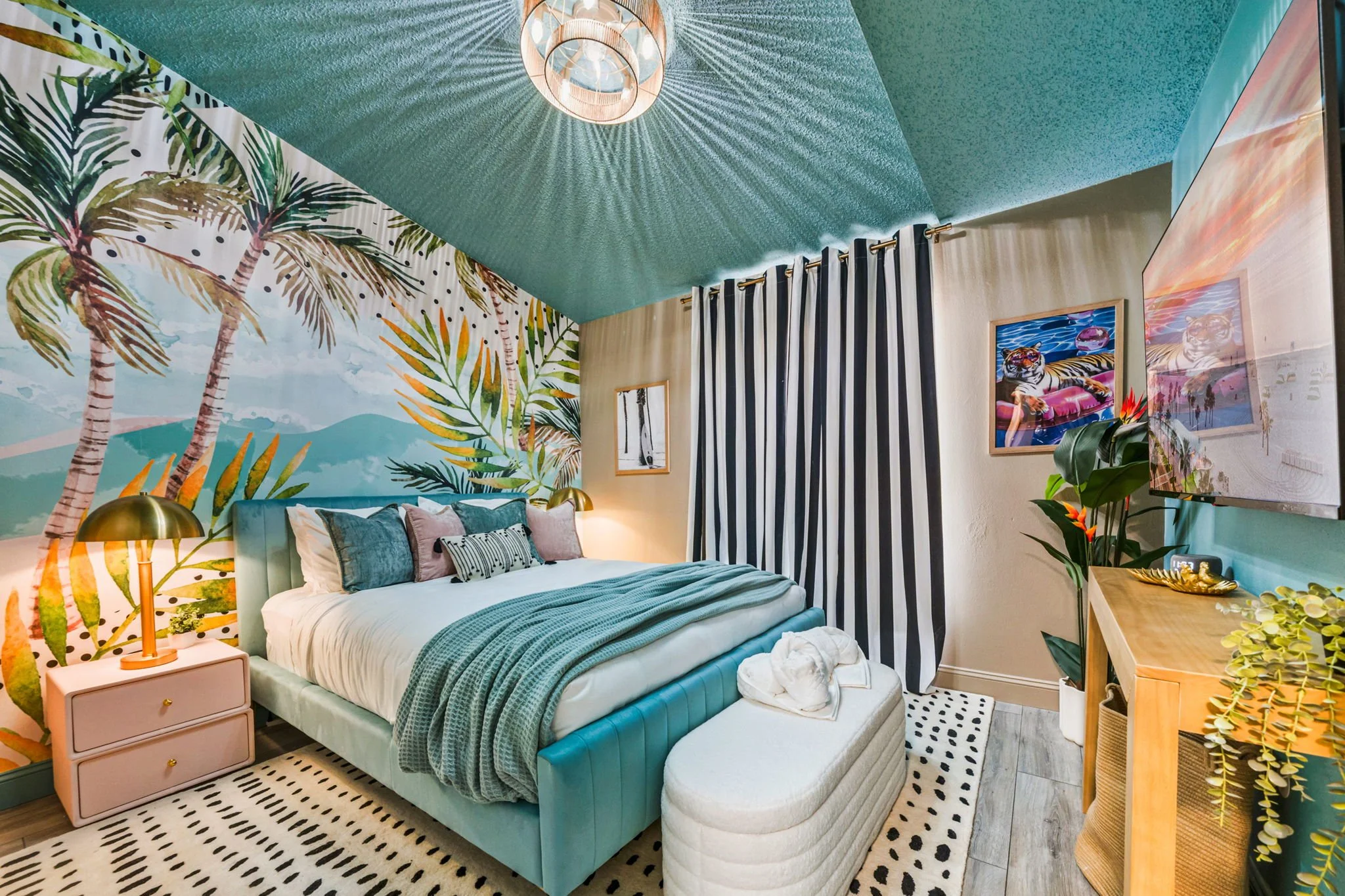 A bedroom with a vibrant tropical theme. The wall behind the bed features a mural of palm trees and mountains. The bed has multiple pillows and a blue headboard. There is a gold bedside lamp on a pink nightstand on the left. A striped black-and-white