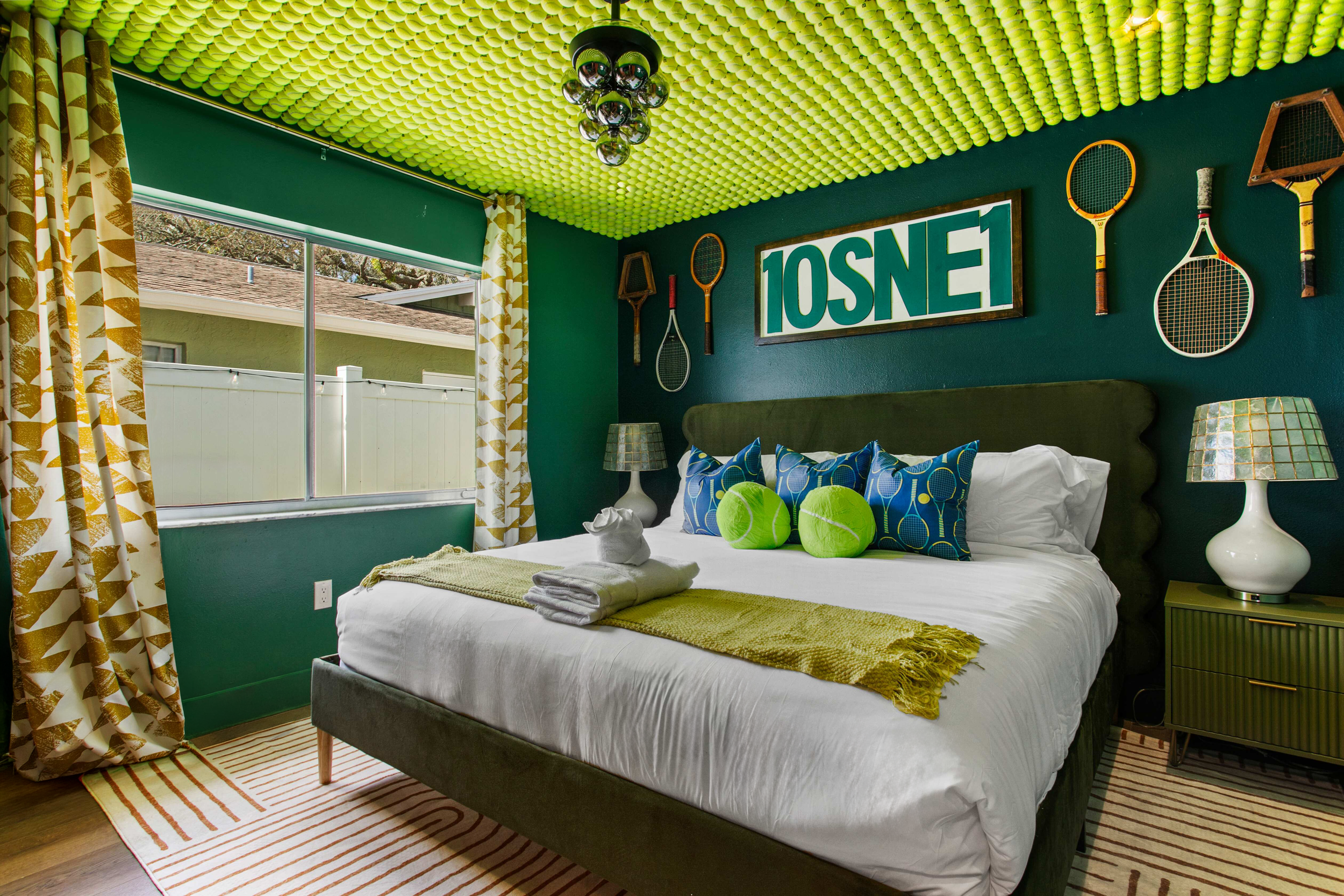 A bedroom with a tennis-themed decor. The room features a green velvet bed with blue and green pillows, a green and gold-striped rug, and matching lamps. The dark green wall behind the bed has tennis rackets and a sign reading "10SNE1." There is a wi