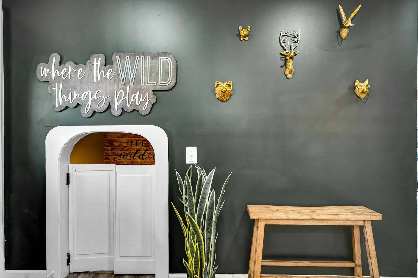 Decorative wall with a sign reading 'where the WILD things play', mounted animal head and face wall mounts, a potted plant, and a wooden table.