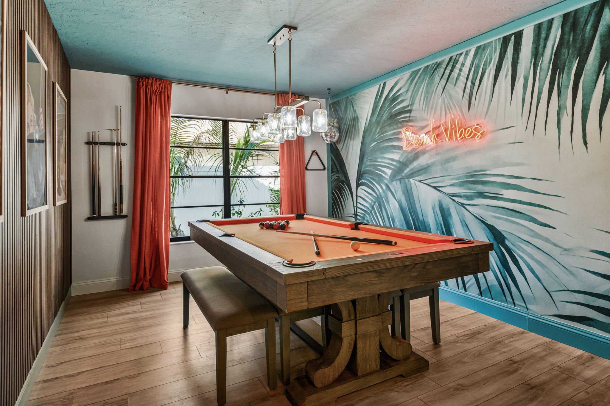 A room with a wooden pool table, tropical-themed mural wall with palm leaves, coral curtains, and a neon sign that says 'Beach Vibes'.