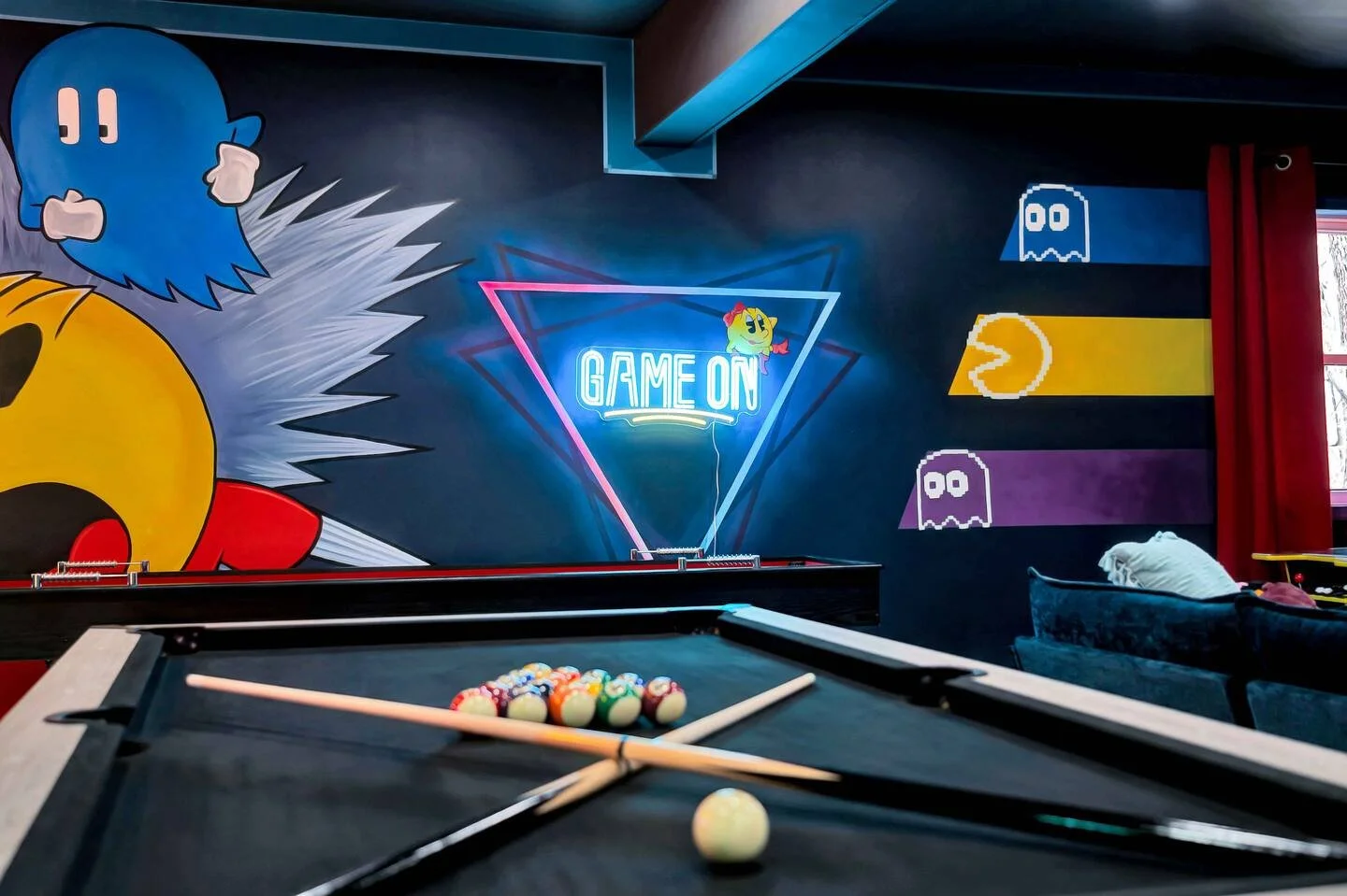 Pool table with billiard balls and cue sticks in a gaming-themed room featuring Pac-Man and arcade game wall art, neon sign that reads 'GAME ON', and pixelated characters on the wall.