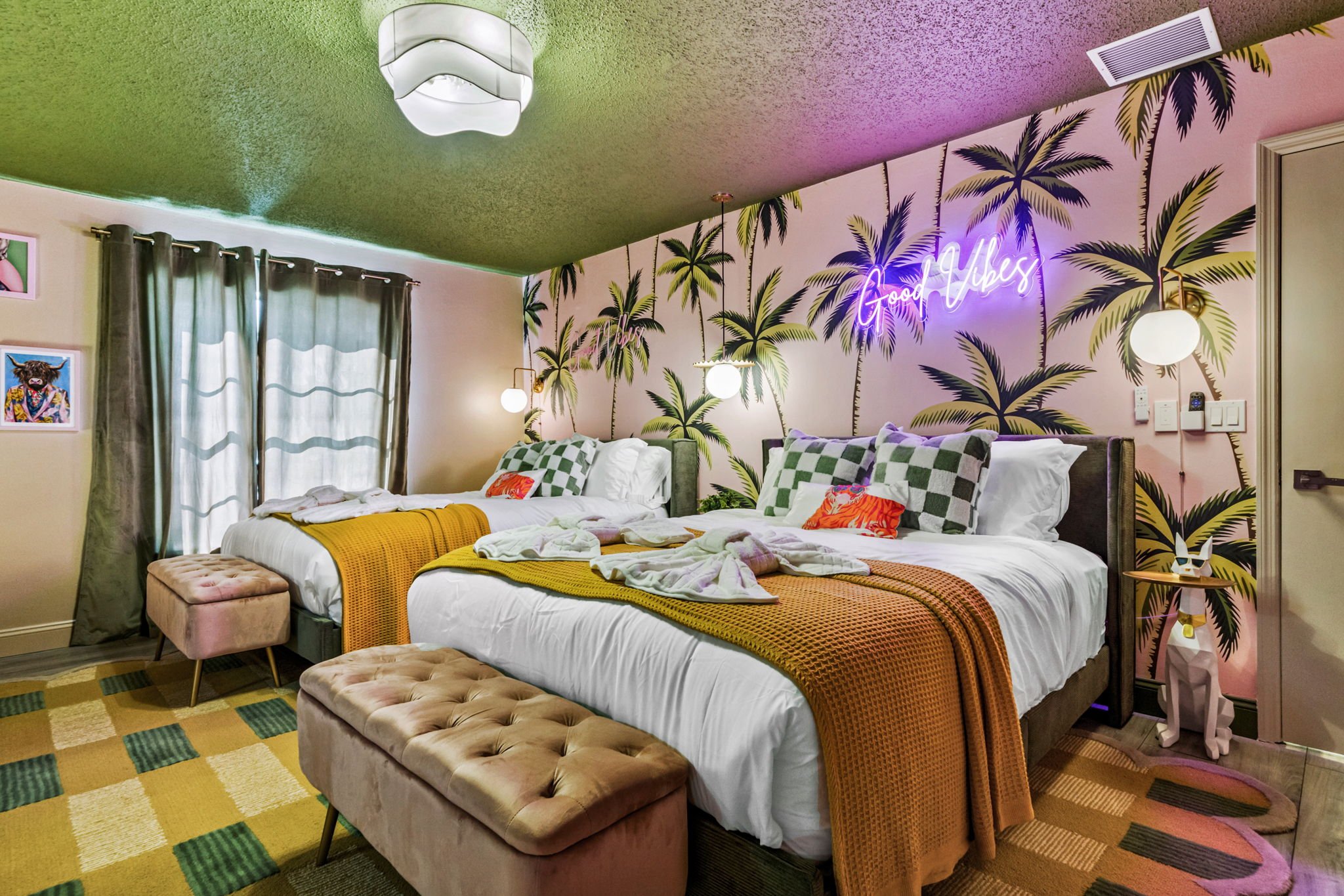 A vibrant hotel room with two queen beds, tropical palm tree wallpaper, and a neon sign reading 'Good Vibes' on the wall. The room features colorful pillows, mustard yellow blankets, a pink ottoman, and a beige tufted bench. Modern lighting and decor