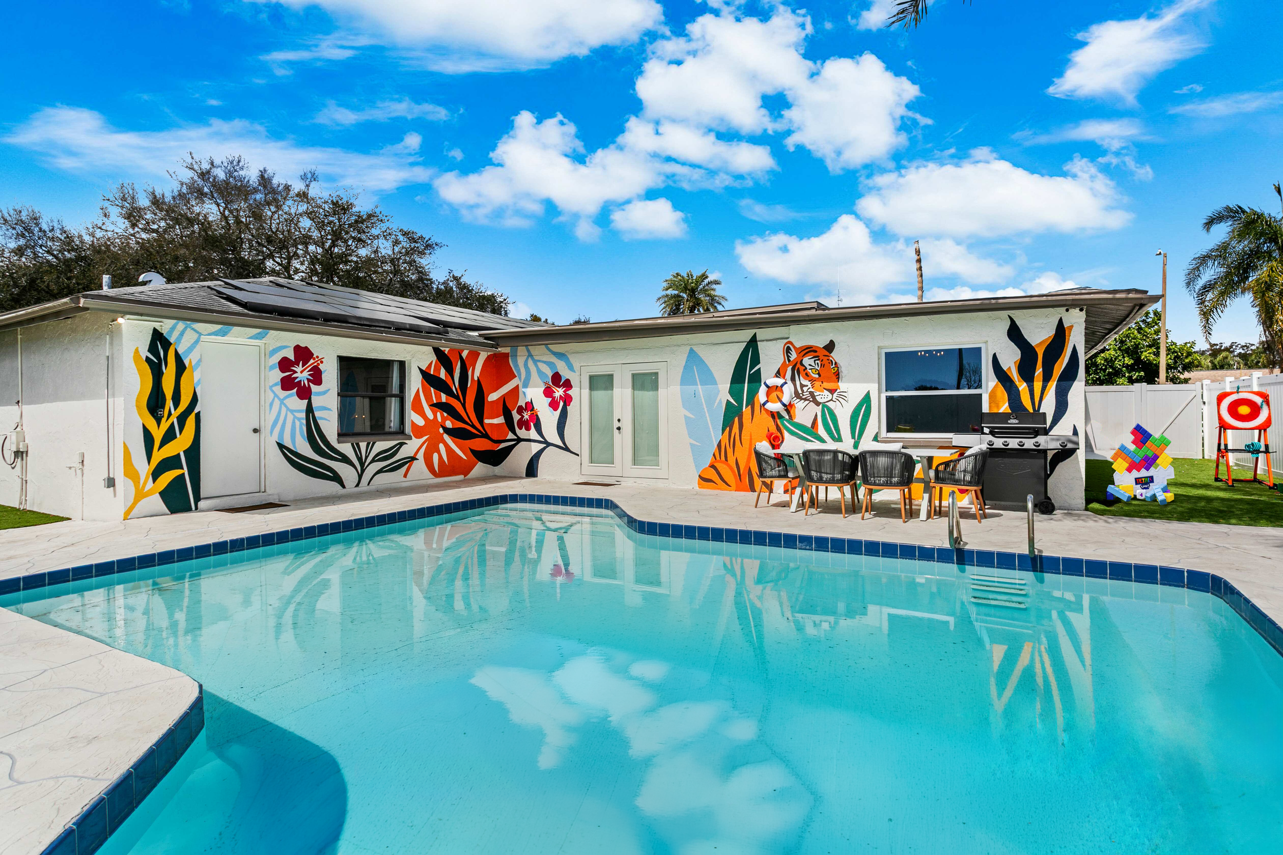 Backyard with pool and house featuring colorful tiger and floral mural wall art, outdoor chairs, barbecue grill, and backyard game equipment under a blue sky with clouds.