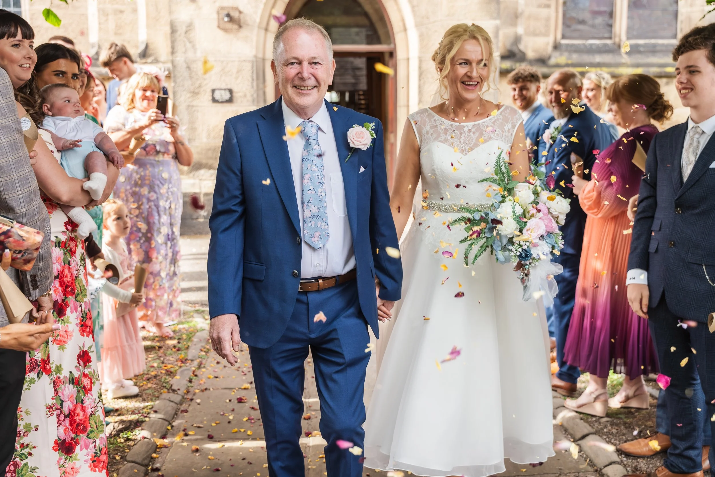 A bride and a man, possibly her father, walking hand in hand as they leave a wedding ceremony. The bride is wearing a white wedding dress and holding a large bouquet of flowers, while the guests are cheering and throwing confetti. The scene is lively and joyful.