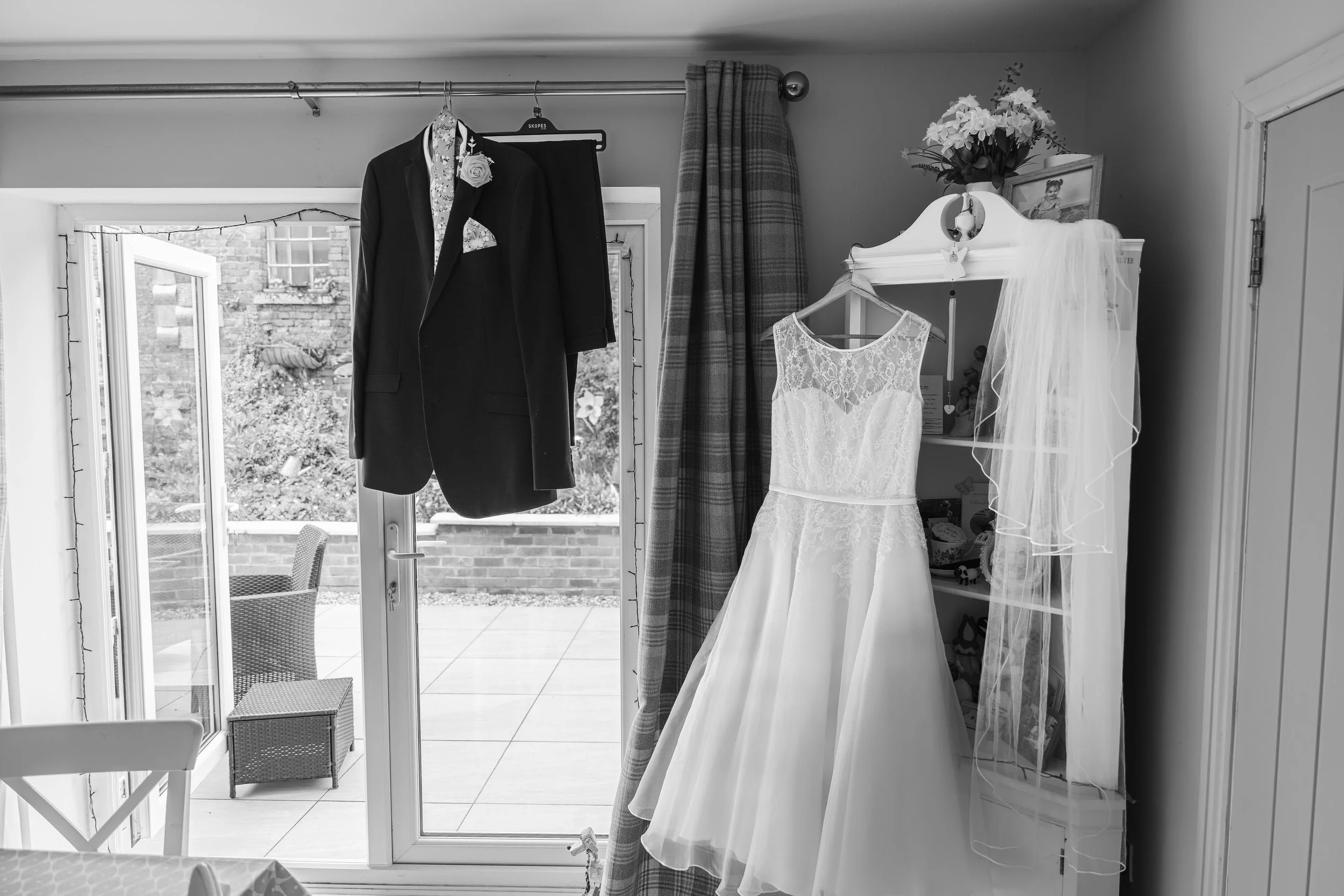 A wedding dress hanging on a shelf, with a veil beside it, and a suit jacket hanging outside the window. The scene appears to be in a room, with a sliding glass door leading to a patio with outdoor furniture.