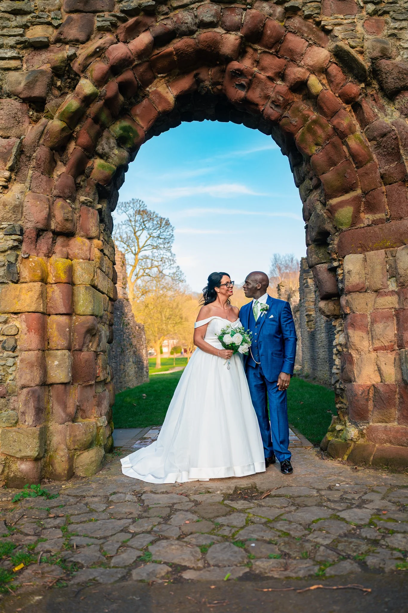 A bride and groom stand under an ancient stone archway, smiling at each other. The bride wears a white wedding dress and holds a bouquet, while the groom wears a blue suit with a white shirt and green tie. The background shows trees and a blue sky.