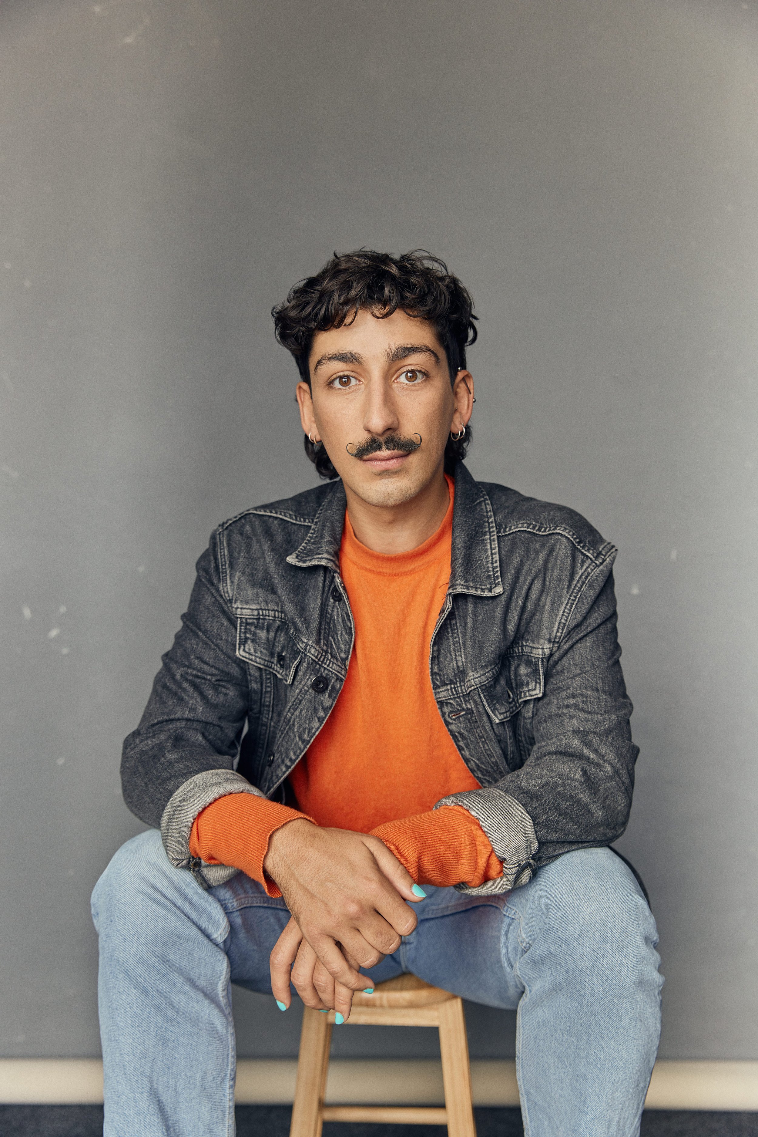 Individual sitting on a stool against a gray background, wearing a denim jacket, orange shirt, light jeans, and painted nails.