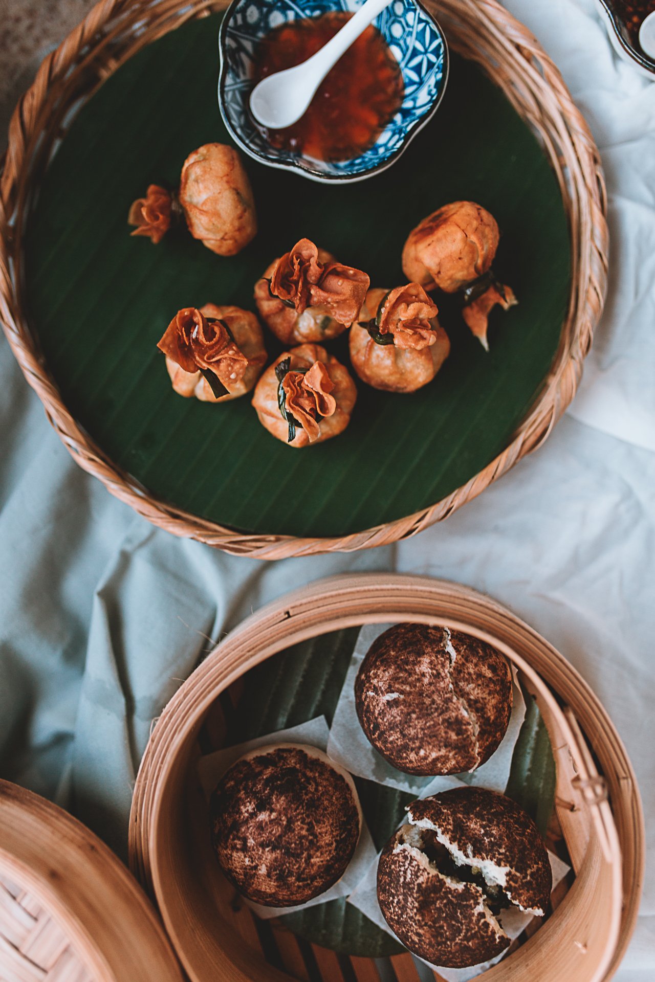 Assorted Thai appetizers on a woven tray with dipping sauce and dumplings, next to buns in a bamboo steamer on a table.
