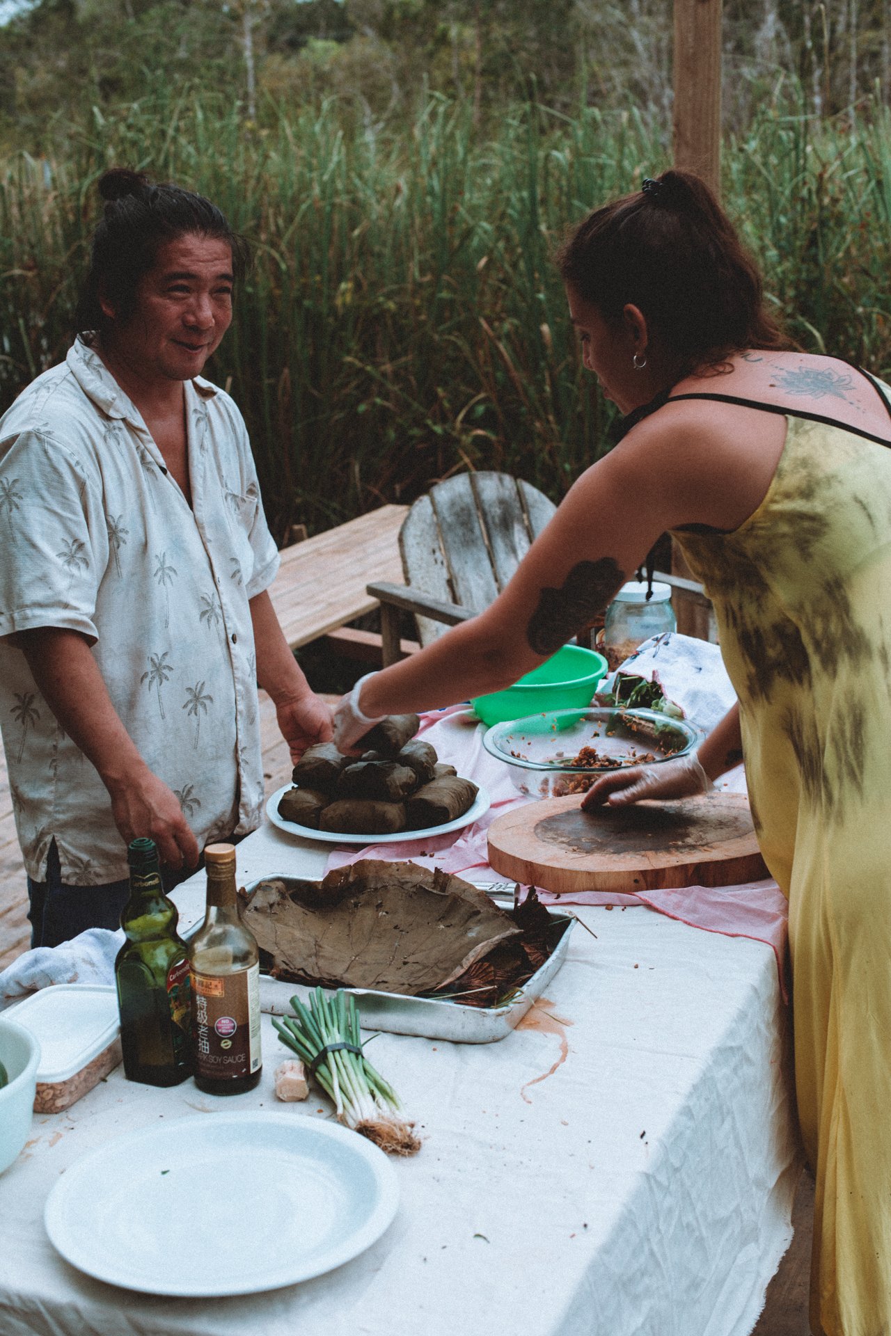 Two people preparing food outdoors, with various dishes and ingredients on a table covered with a white cloth.
