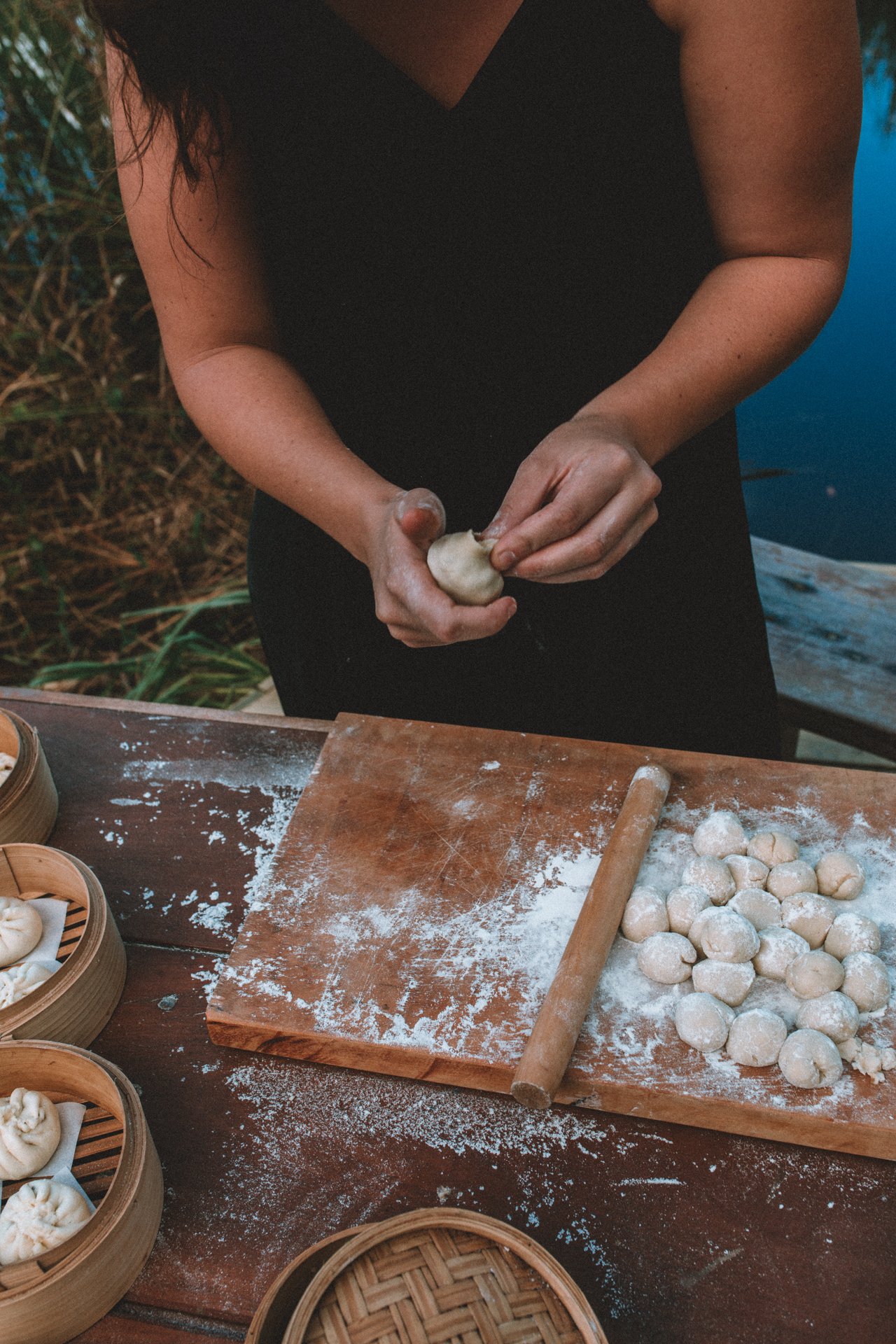 Person making dumplings on a flour-dusted wooden surface with bamboo steamers.