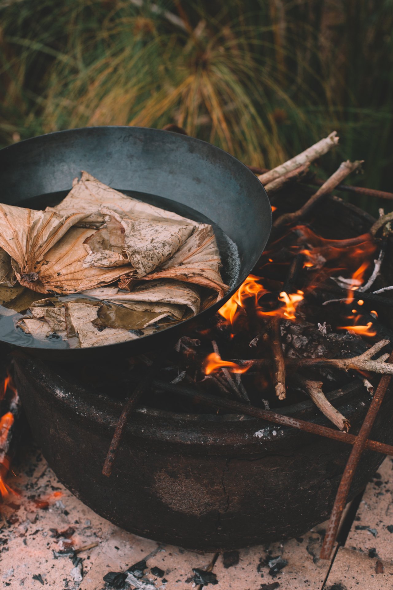 A pot of water with dried leaves over a burning fire, surrounded by wood and metal bars, outdoors.