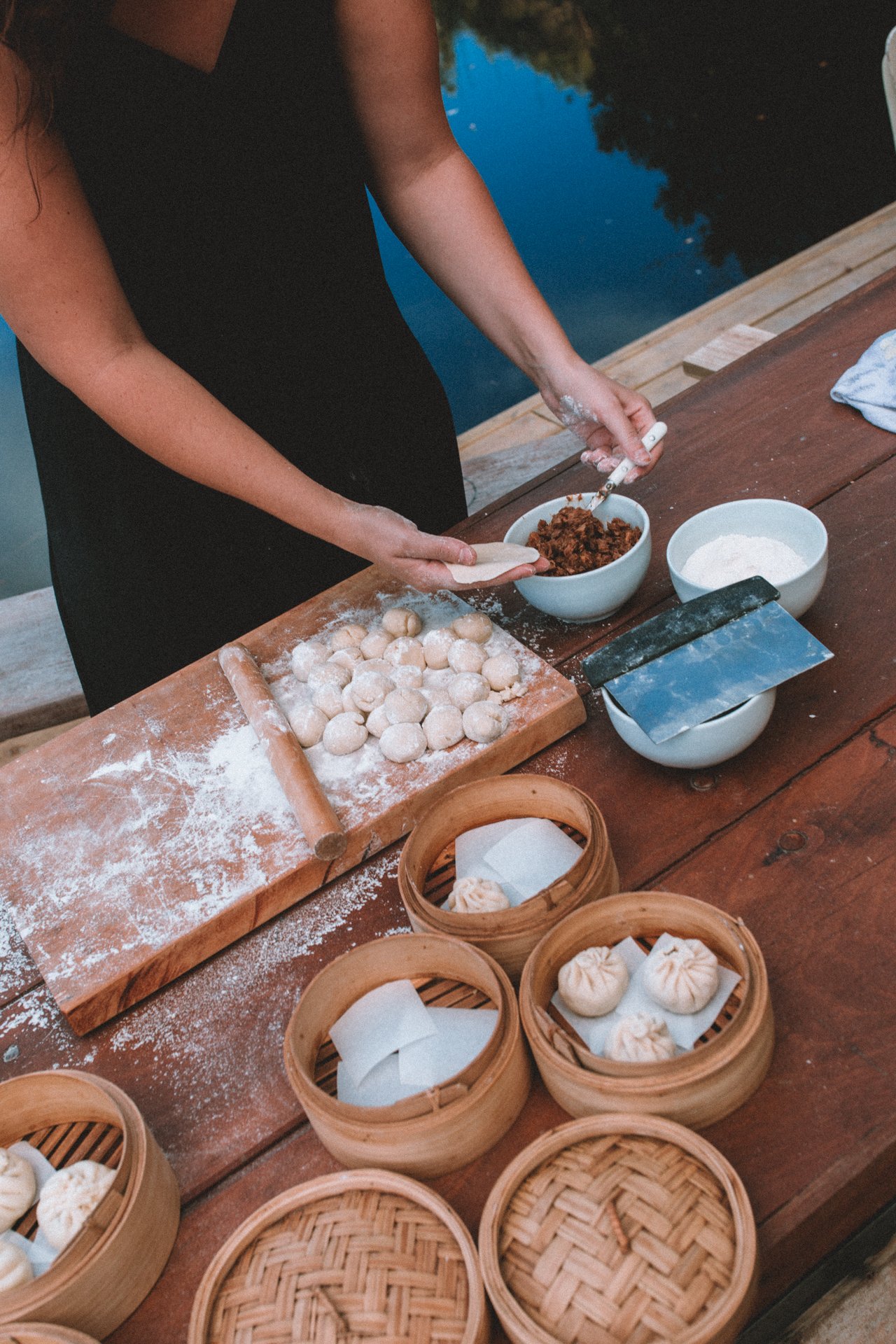 Person preparing dumplings with dough and filling on a wooden table, surrounded by bamboo steamers and bowls containing ingredients.