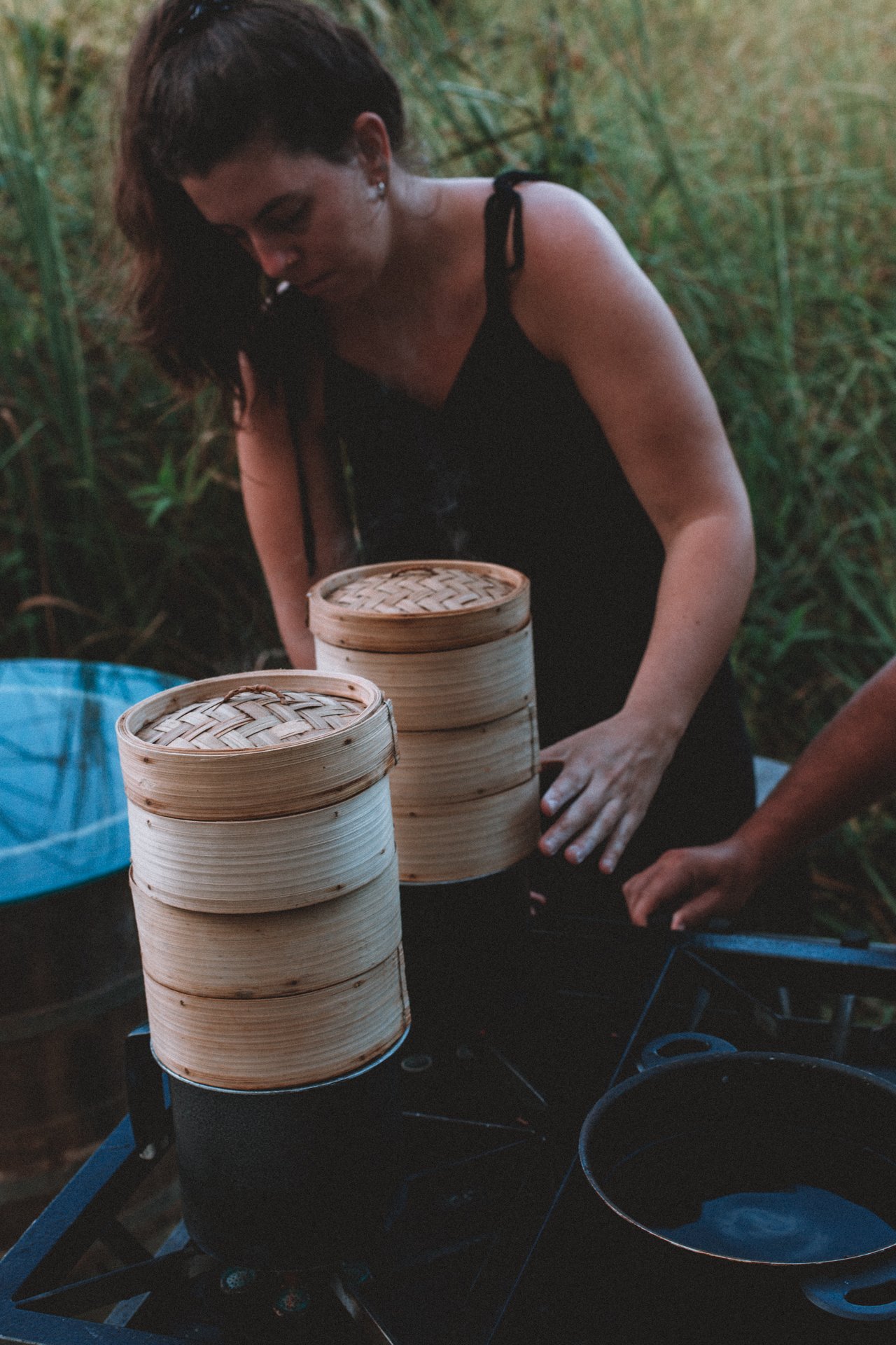 Woman cooking outdoors with bamboo steamers on a portable stove.