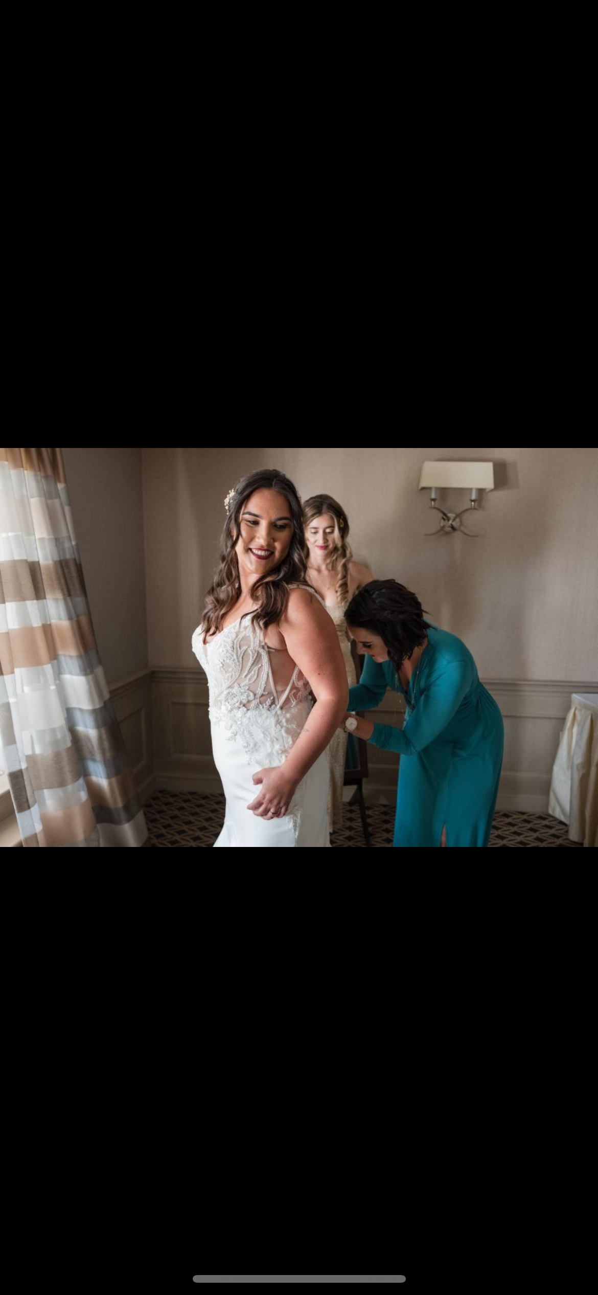 Bride smiling while getting ready, two women assisting with her dress in a room with patterned curtains and wall lamp.