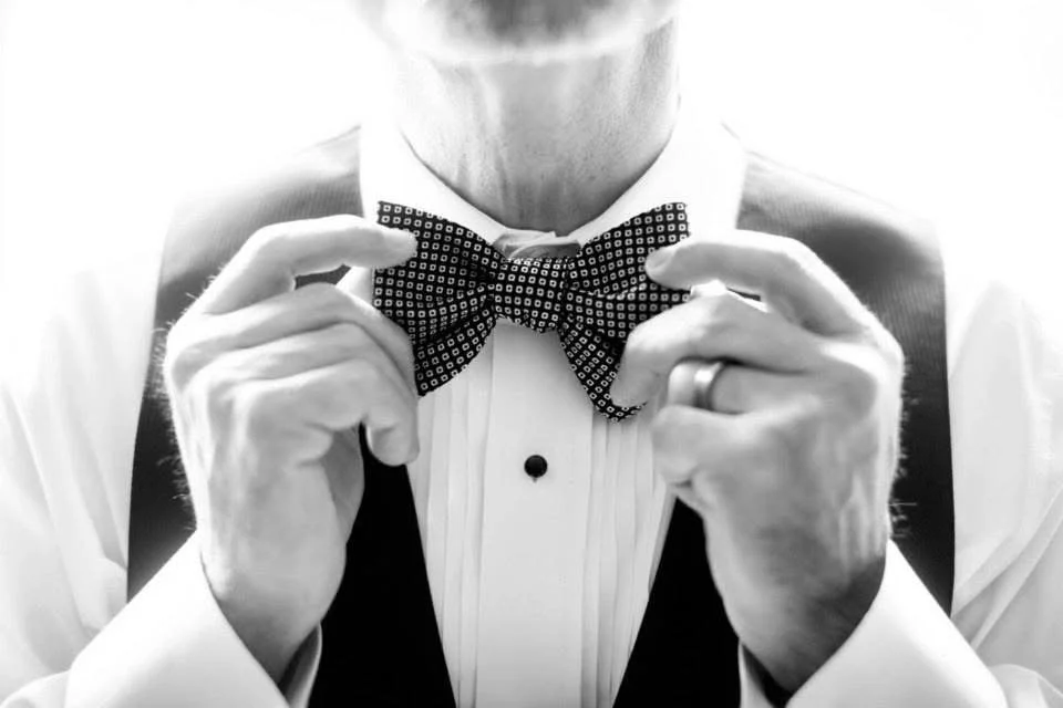 Man adjusting a patterned bow tie, wearing a tuxedo with a vest, in black and white.