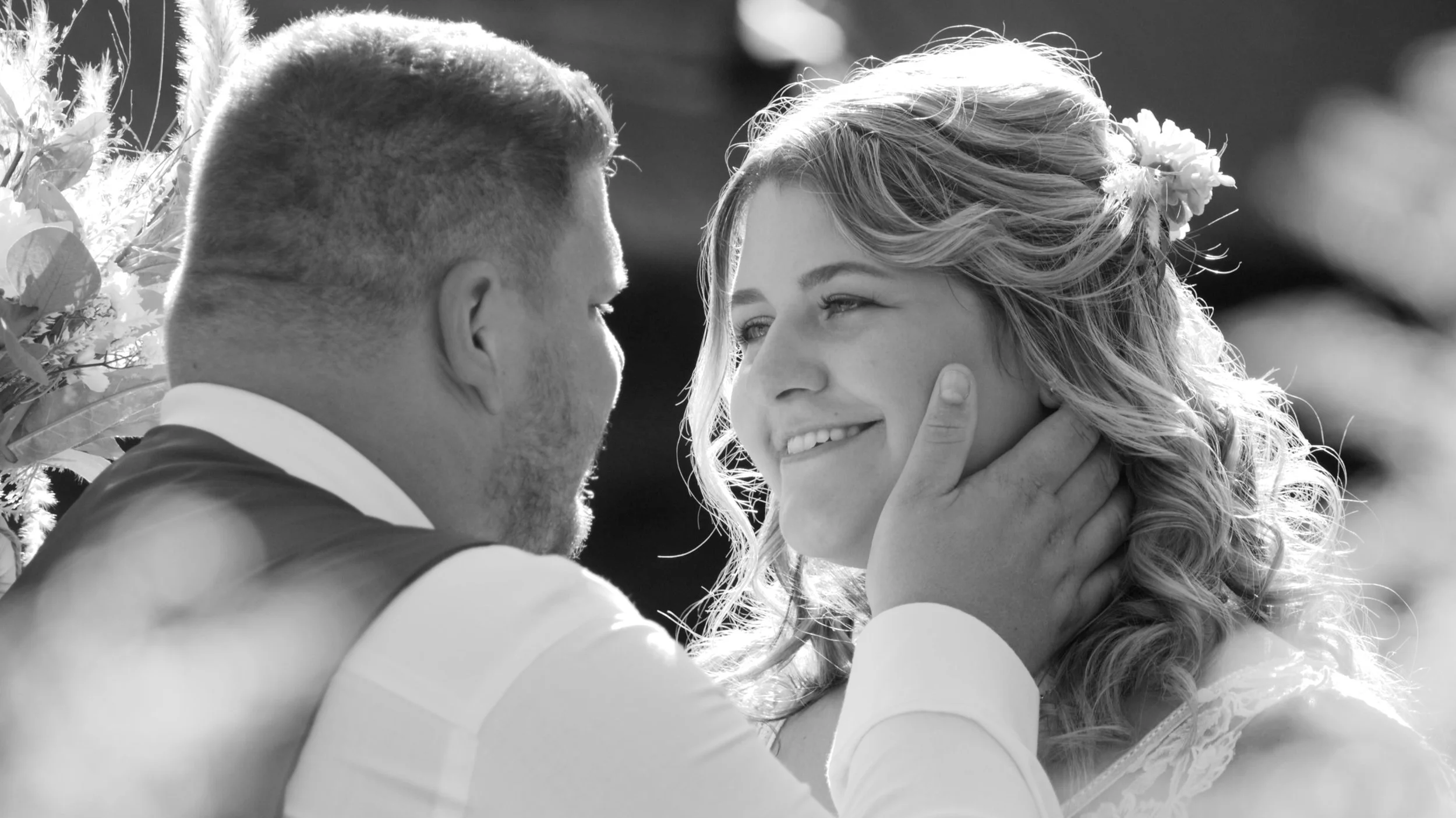 A black-and-white photograph of a couple with their faces close, the woman smiling with her hand on the man's neck, outdoors with natural light.