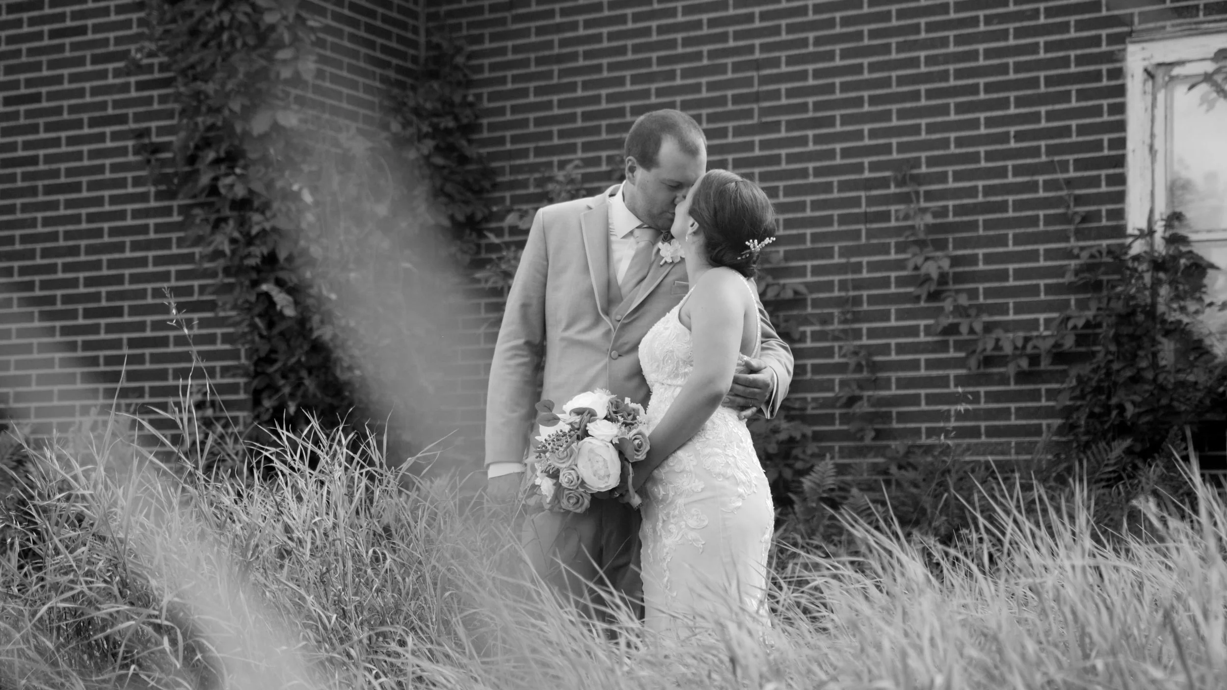 A bride and groom sharing a kiss outdoors, with the bride holding a bouquet of roses and wearing a lace wedding dress, and the groom dressed in a suit, standing in front of a brick wall surrounded by tall grass and plants.