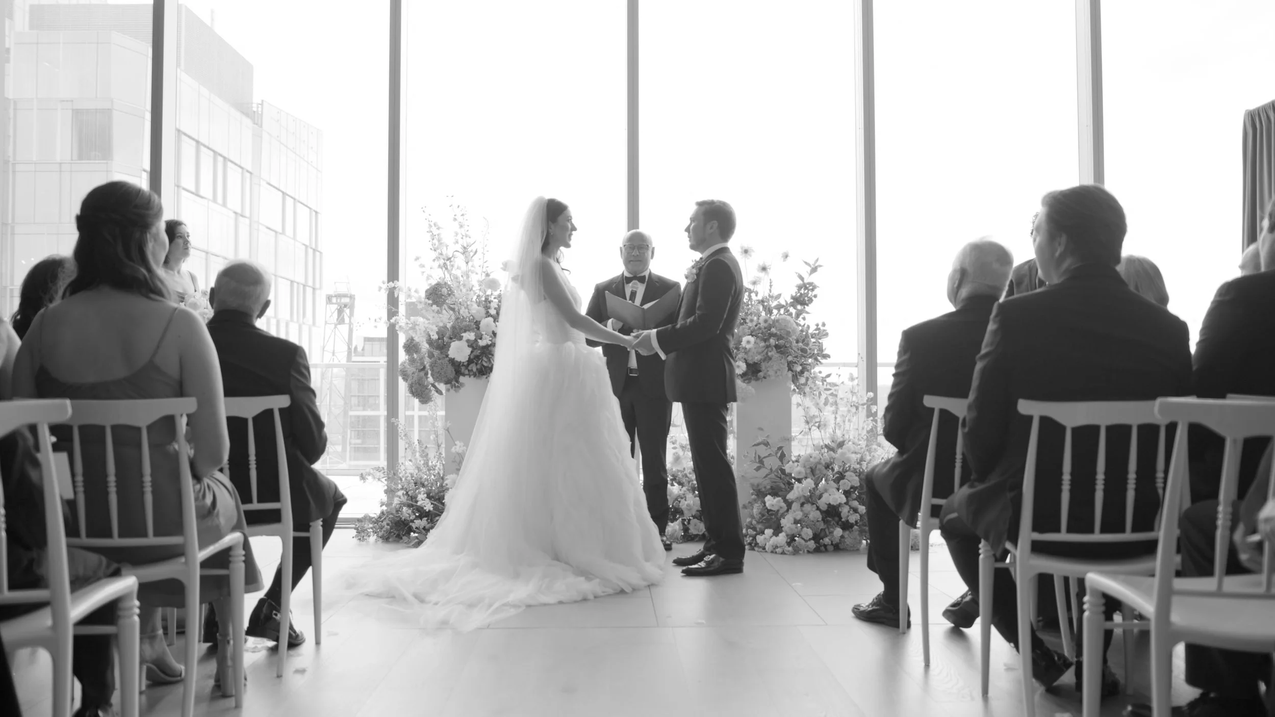 A wedding ceremony with a bride and groom holding hands, standing in front of a minister, with guests seated on either side and large windows in the background.