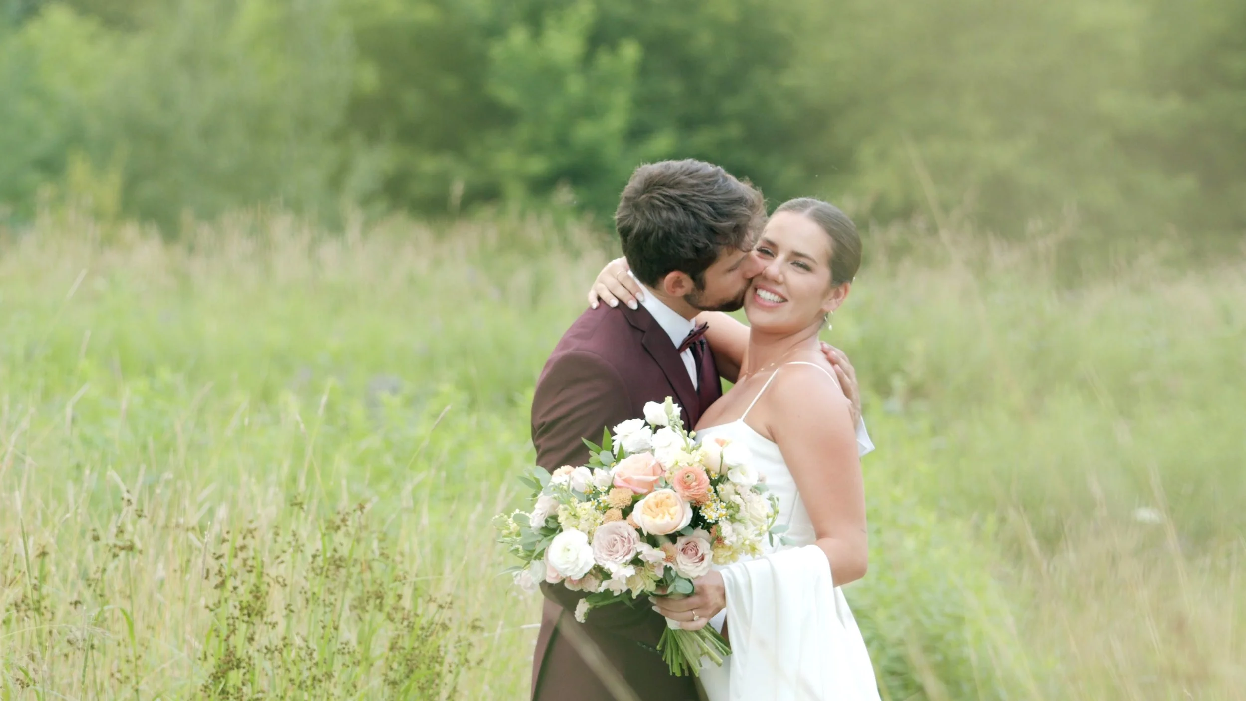 A newlywed couple embraces in a grassy field, the man kissing the woman's cheek as she holds a bouquet of pink and white roses, both smiling.