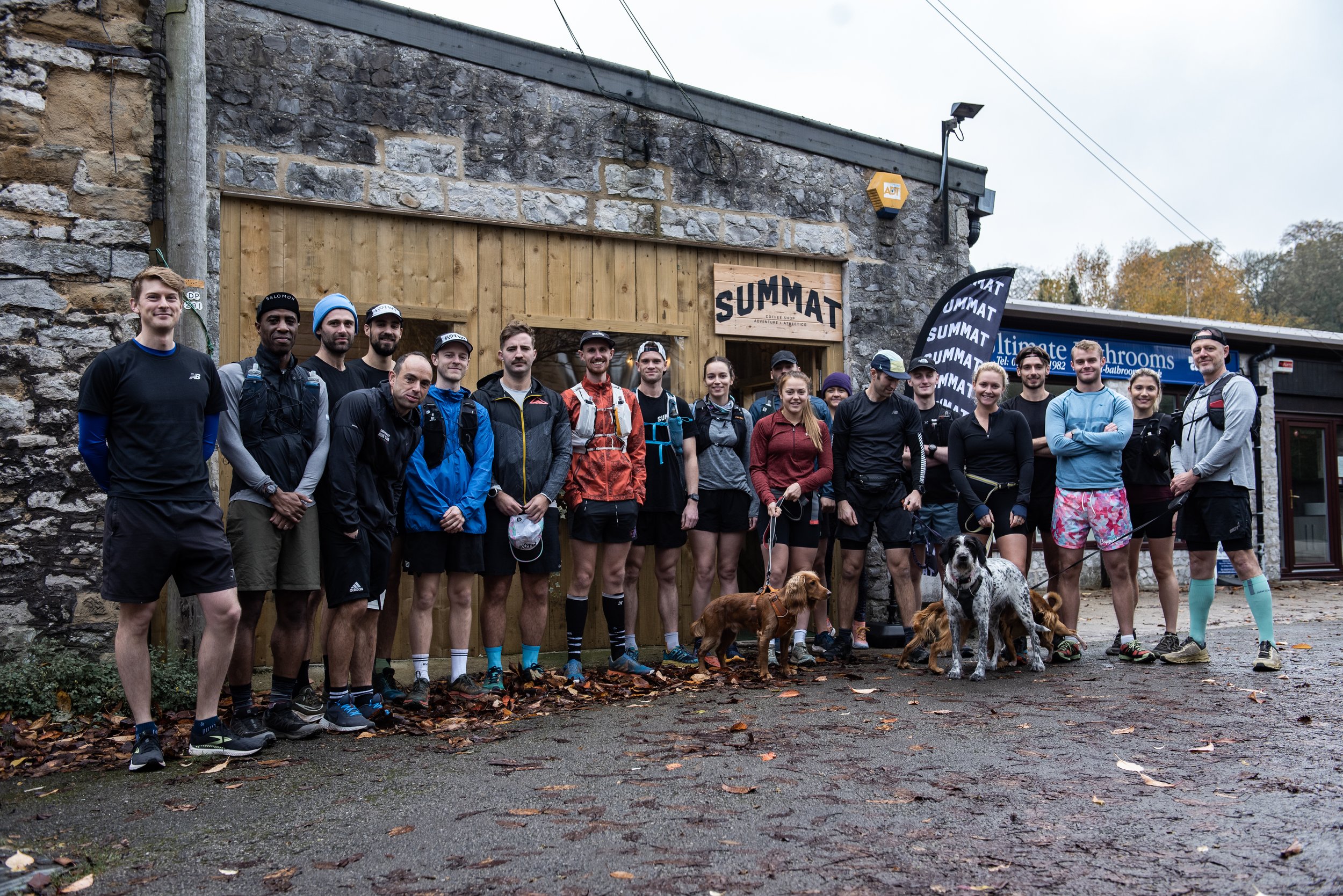 Group of runners with dogs standing outside a stone building labeled 'Summat,' wearing casual athletic clothing, some holding dog leashes, on a paved area covered with autumn leaves.