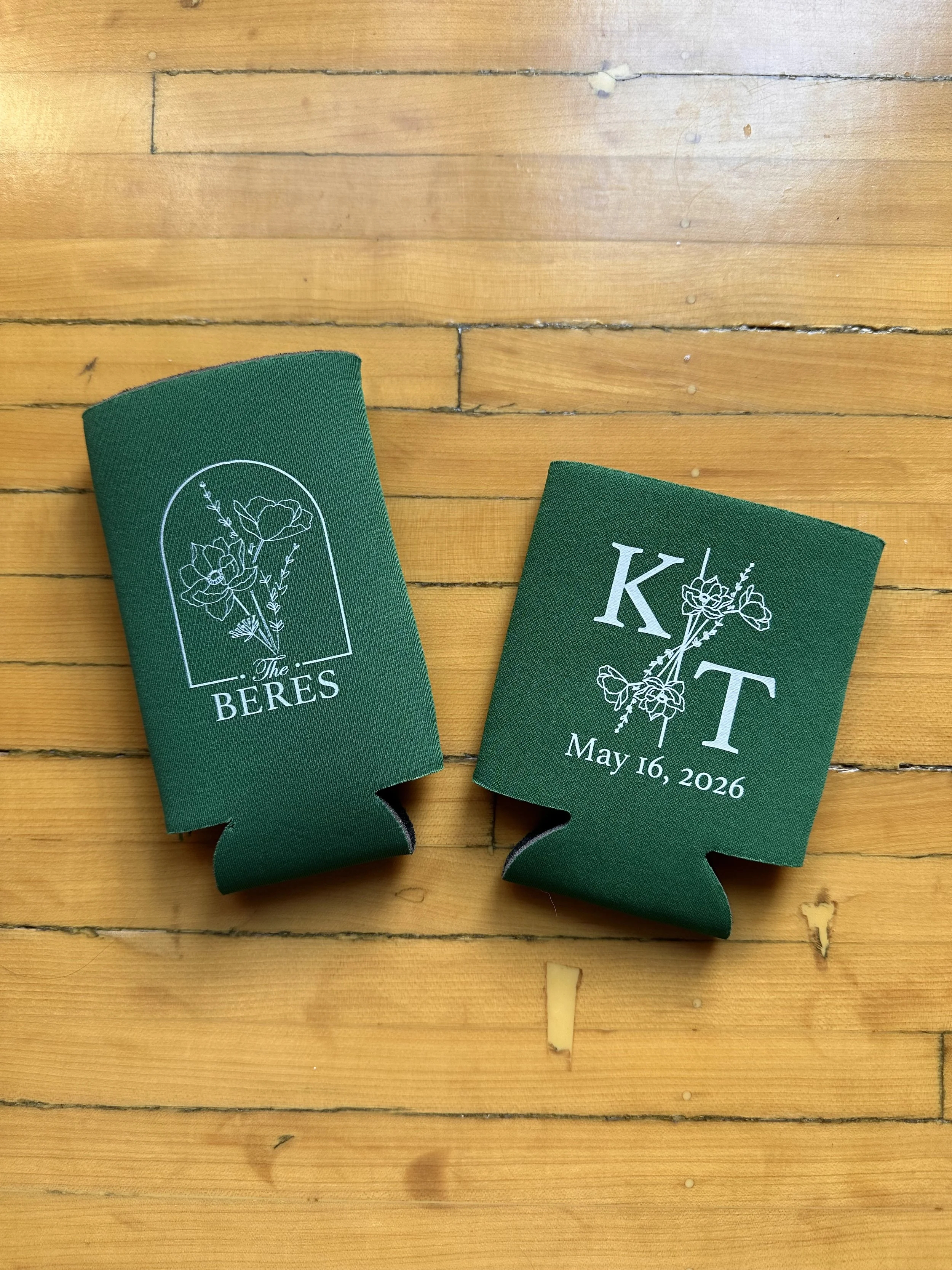 Two green ribbon bookmarks, one with a floral design and the words "The BERES", and the other with the letter "K" and a floral design, on a wooden surface.