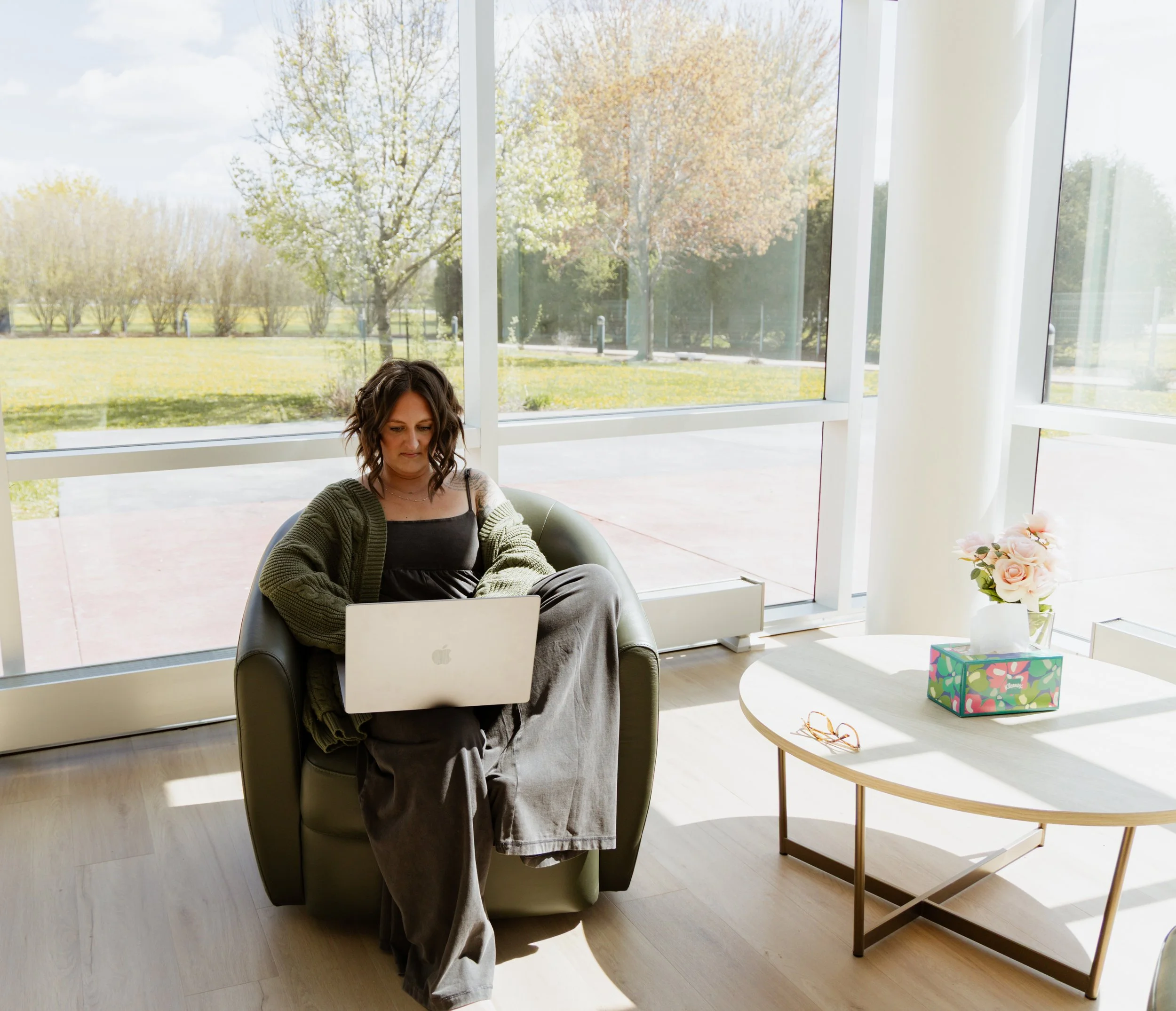 Woman sitting in a modern room with large windows and an outdoor view, working on a laptop at a round table with a floral gift box and sunglasses.