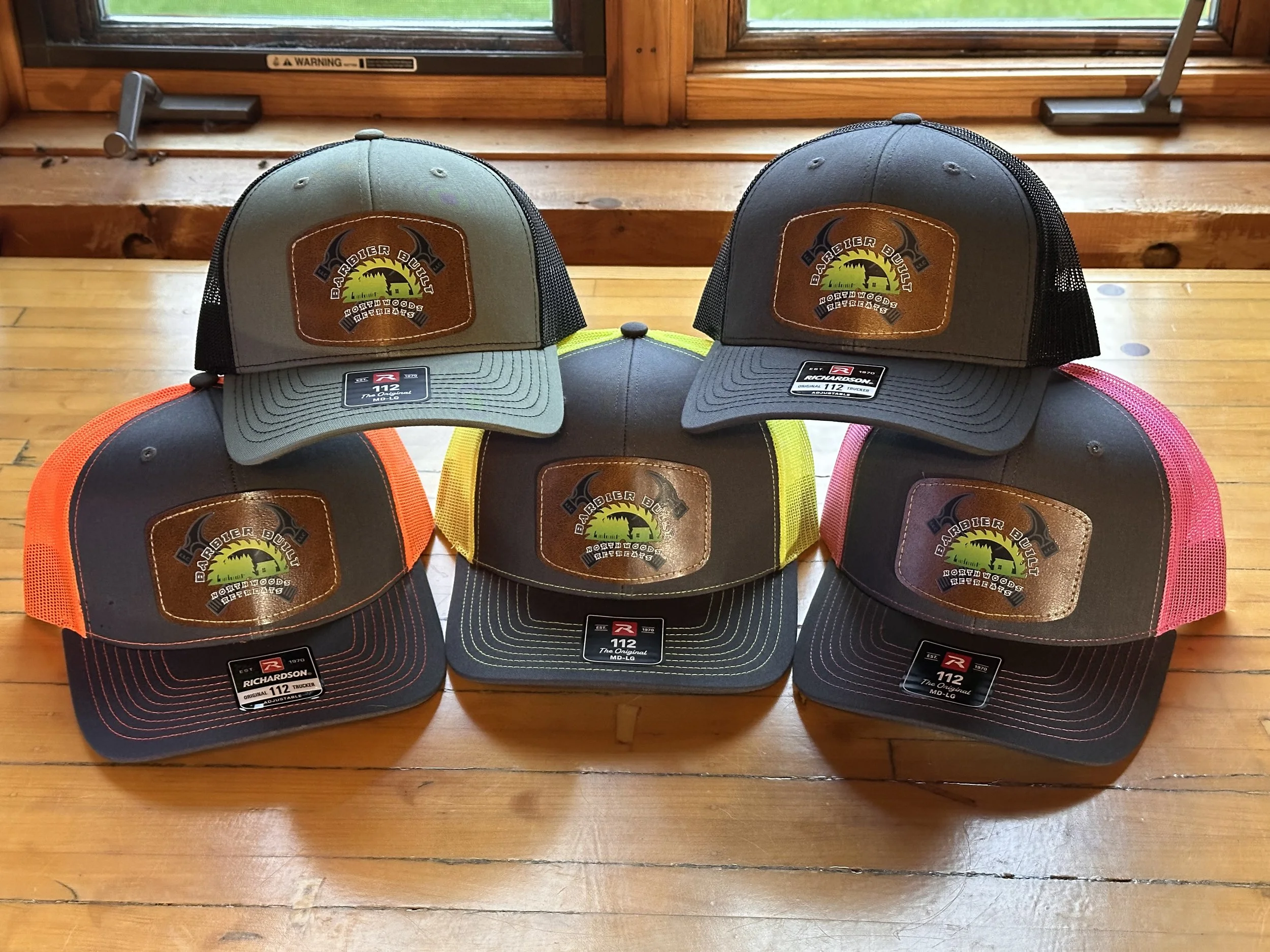 Six trucker hats with different colors on a wooden table, each featuring a patch with a logo that reads "Barbier Built Northwoods Retreats" and a stylized graphic of a house and trees.