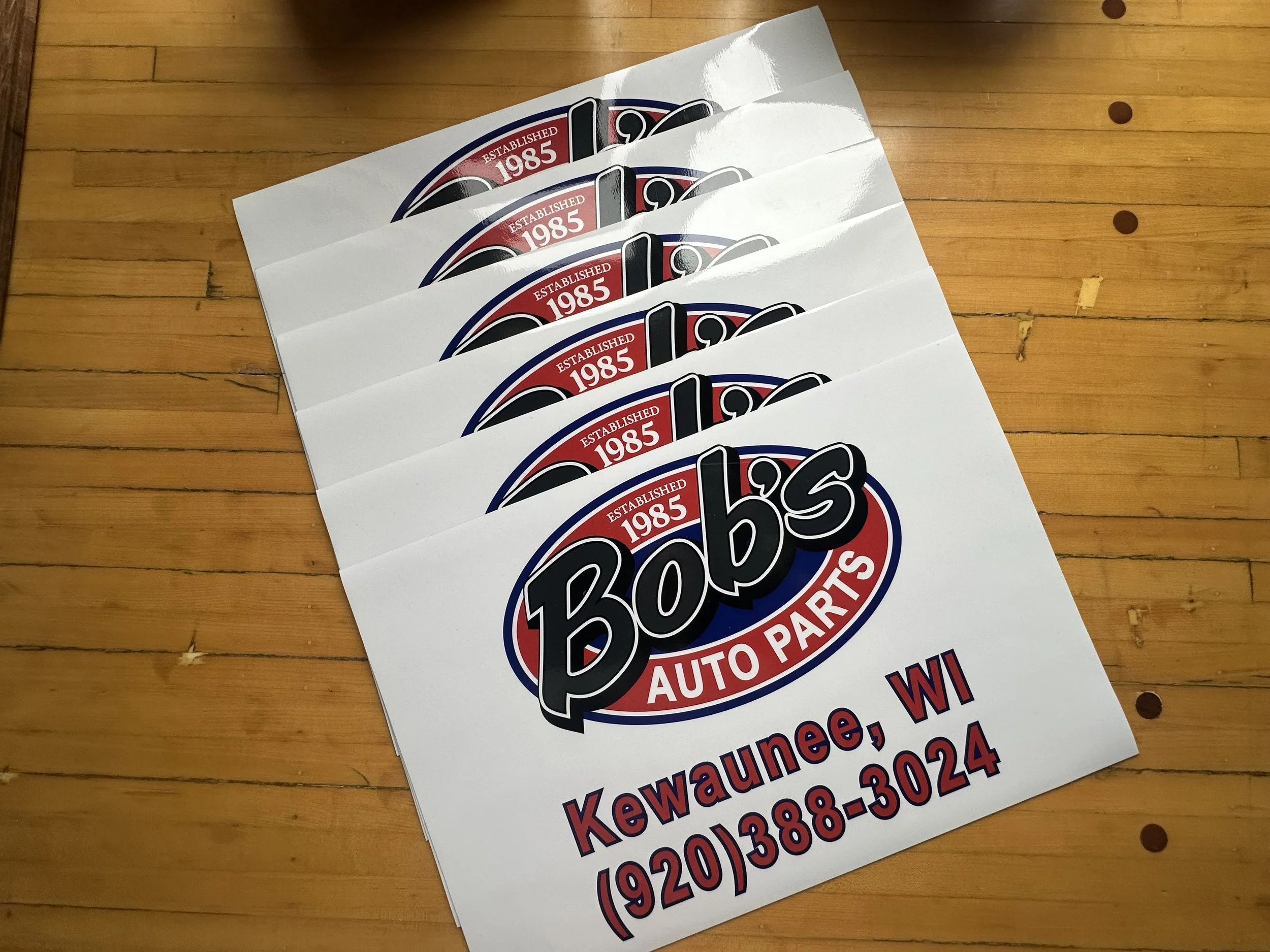 Promotional signs for auto parts store with the logo, location in Wisconsin, and contact information on a wooden surface.