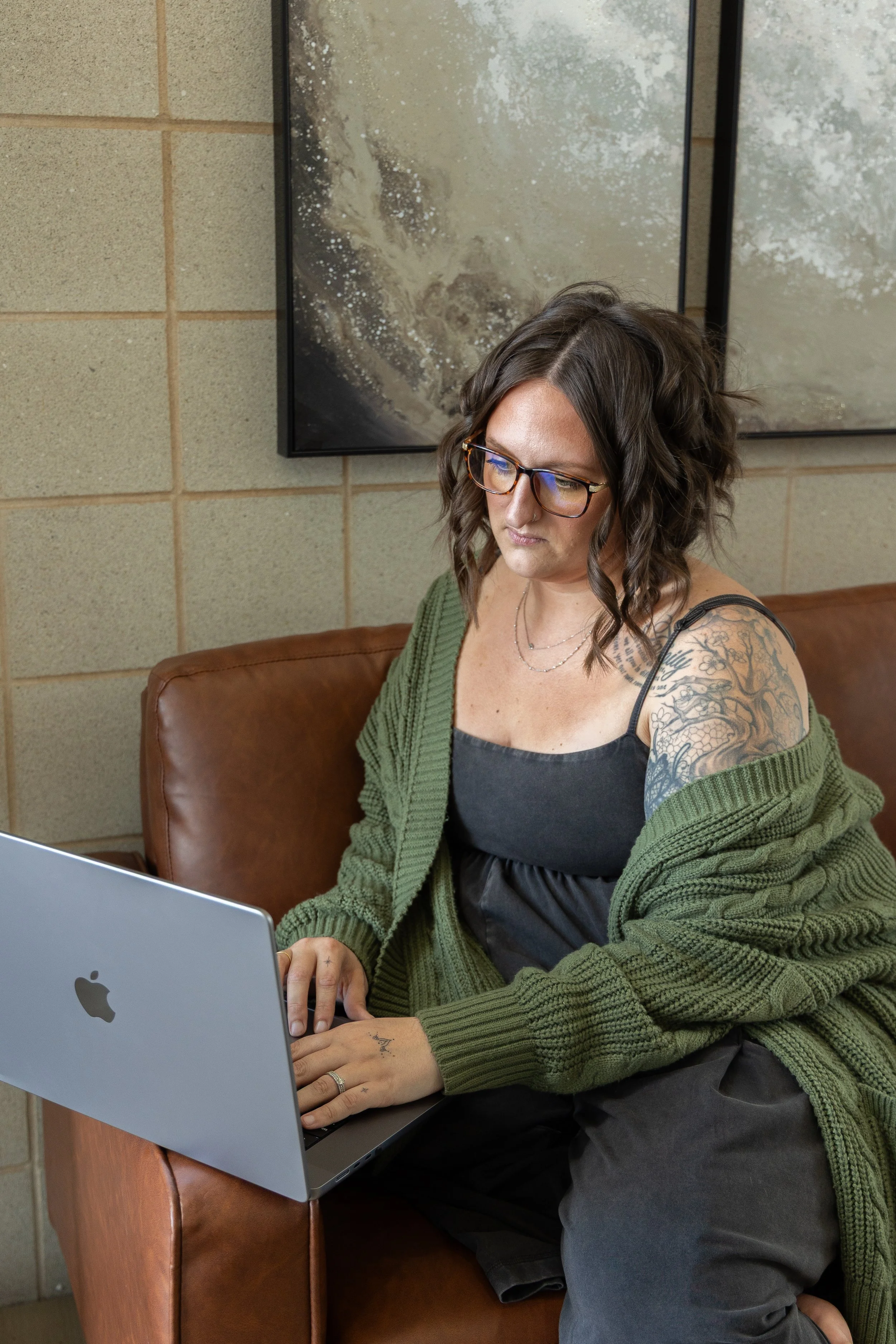 A woman with glasses and tattoos sitting on a brown leather couch working on a silver MacBook laptop, wearing a black dress and green cardigan, with a textured beige wall and abstract art behind her.
