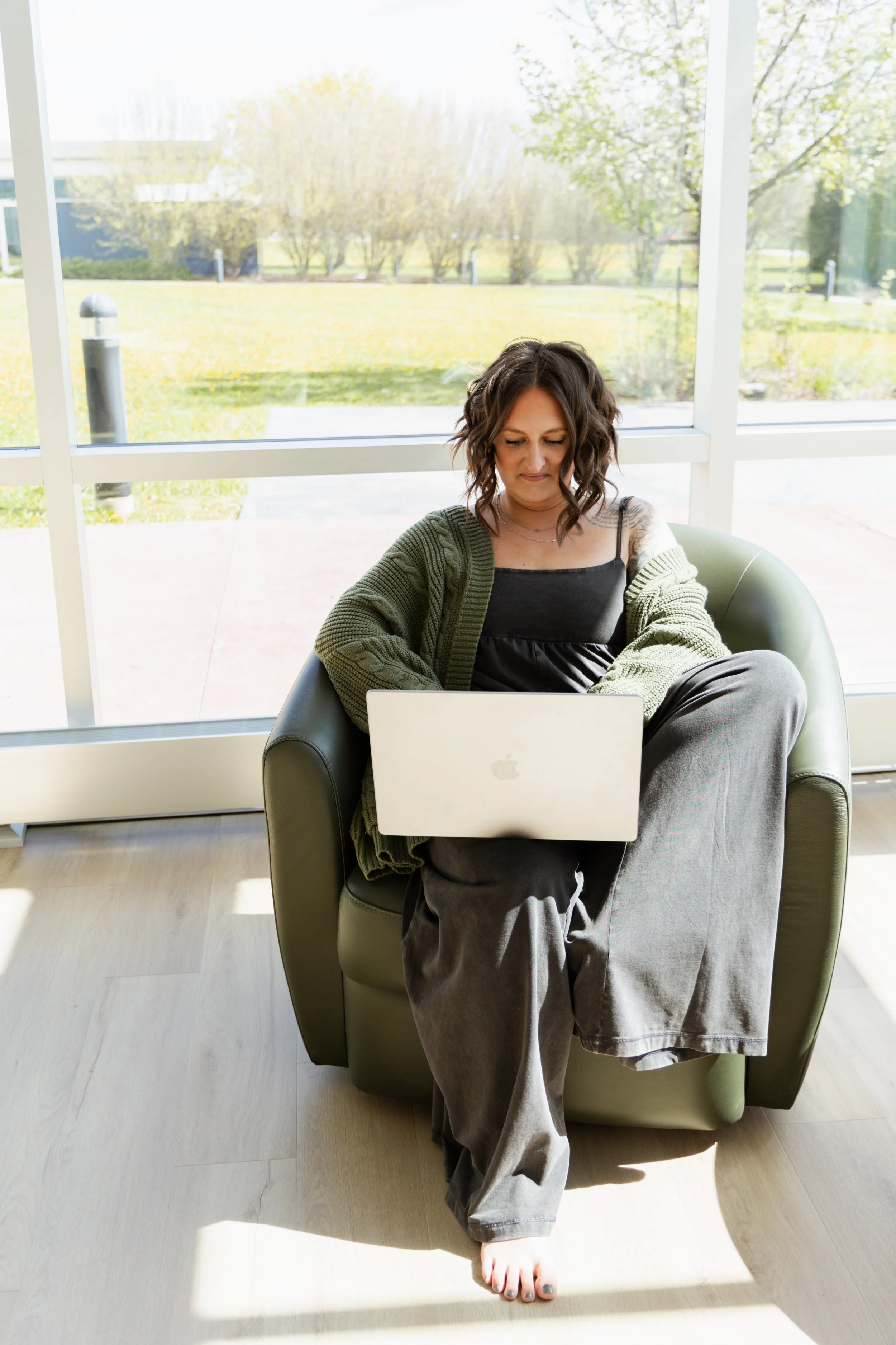 A woman sitting on a green armchair with a laptop on her lap, barefoot, inside a room with large windows showing a sunny outdoor landscape with trees and grass.
