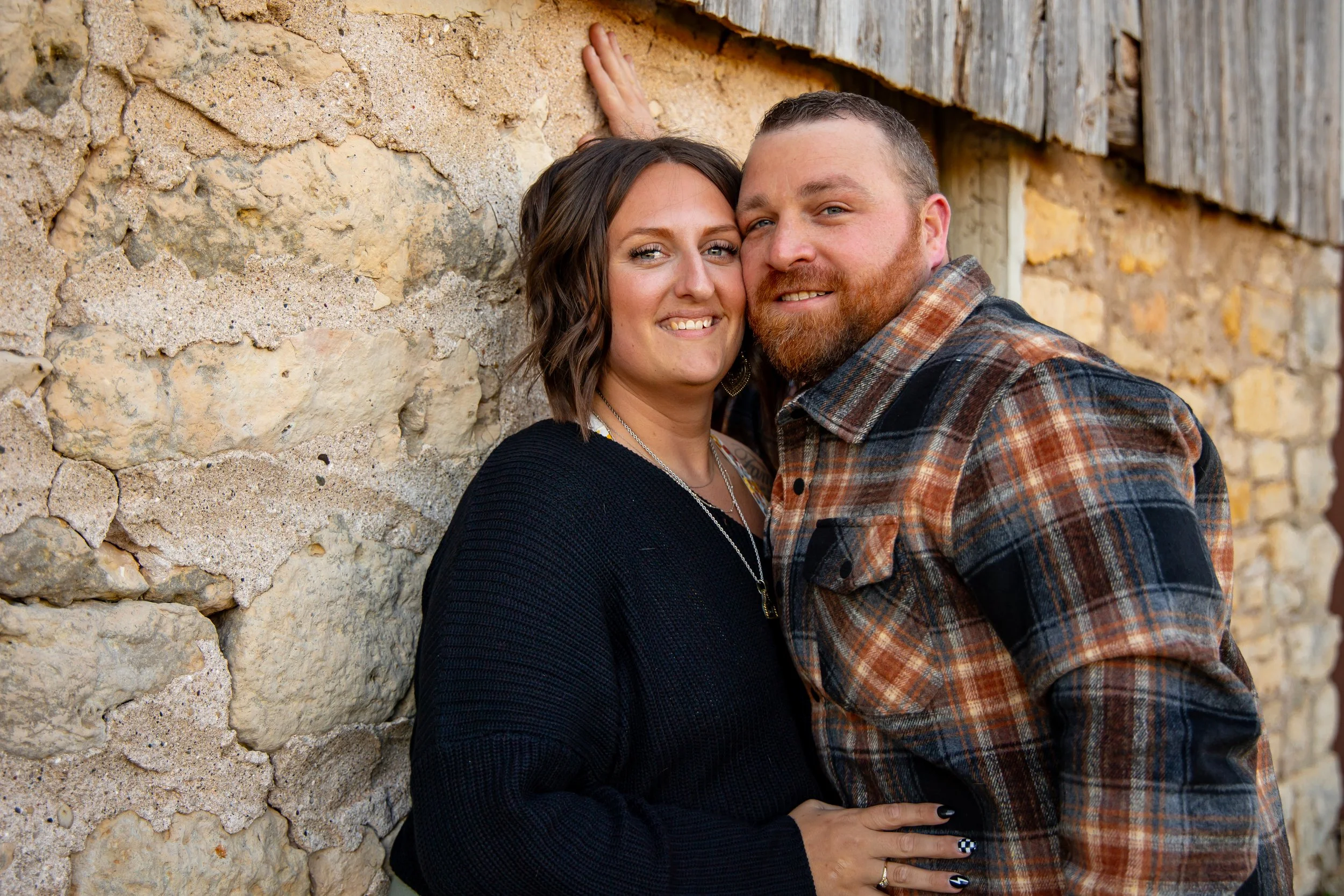 A smiling couple, a woman with dark wavy hair and a man with a beard, embracing in front of a textured stone wall with wooden beams.