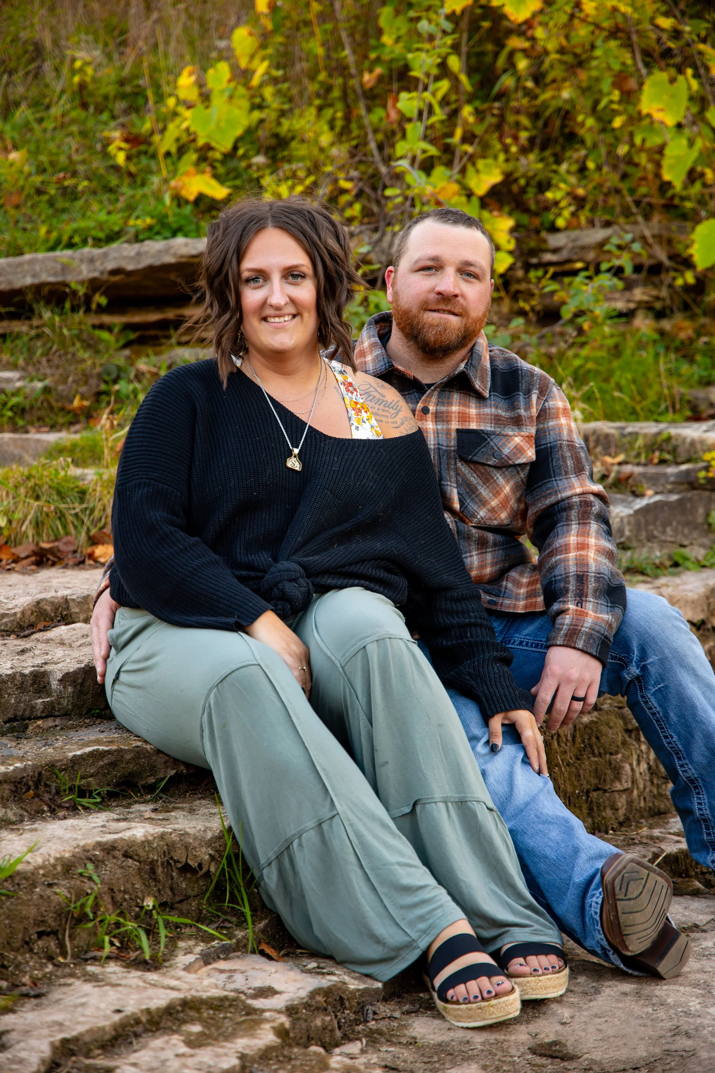 A couple sitting on stone steps outdoors during fall, smiling at the camera. The woman has shoulder-length brown hair, is wearing a black sweater, light-colored pants, and sandals. The man has short hair and a beard, is wearing a plaid shirt and jeans. The background is filled with green and yellow foliage.