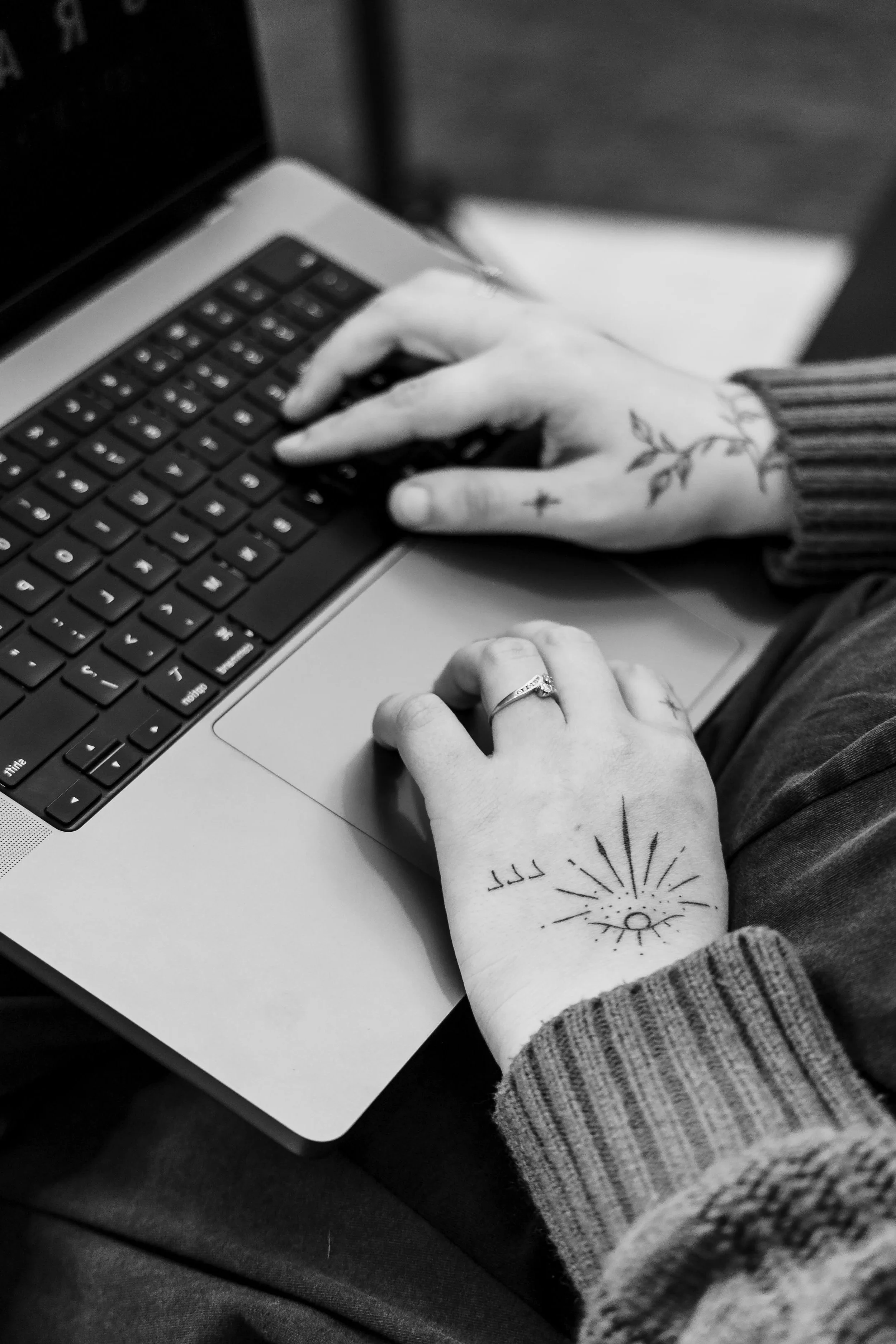 A person's hand with tattoos on the wrist, using a laptop keyboard. The person wears a striped long sleeve shirt and a ring on the ring finger.
