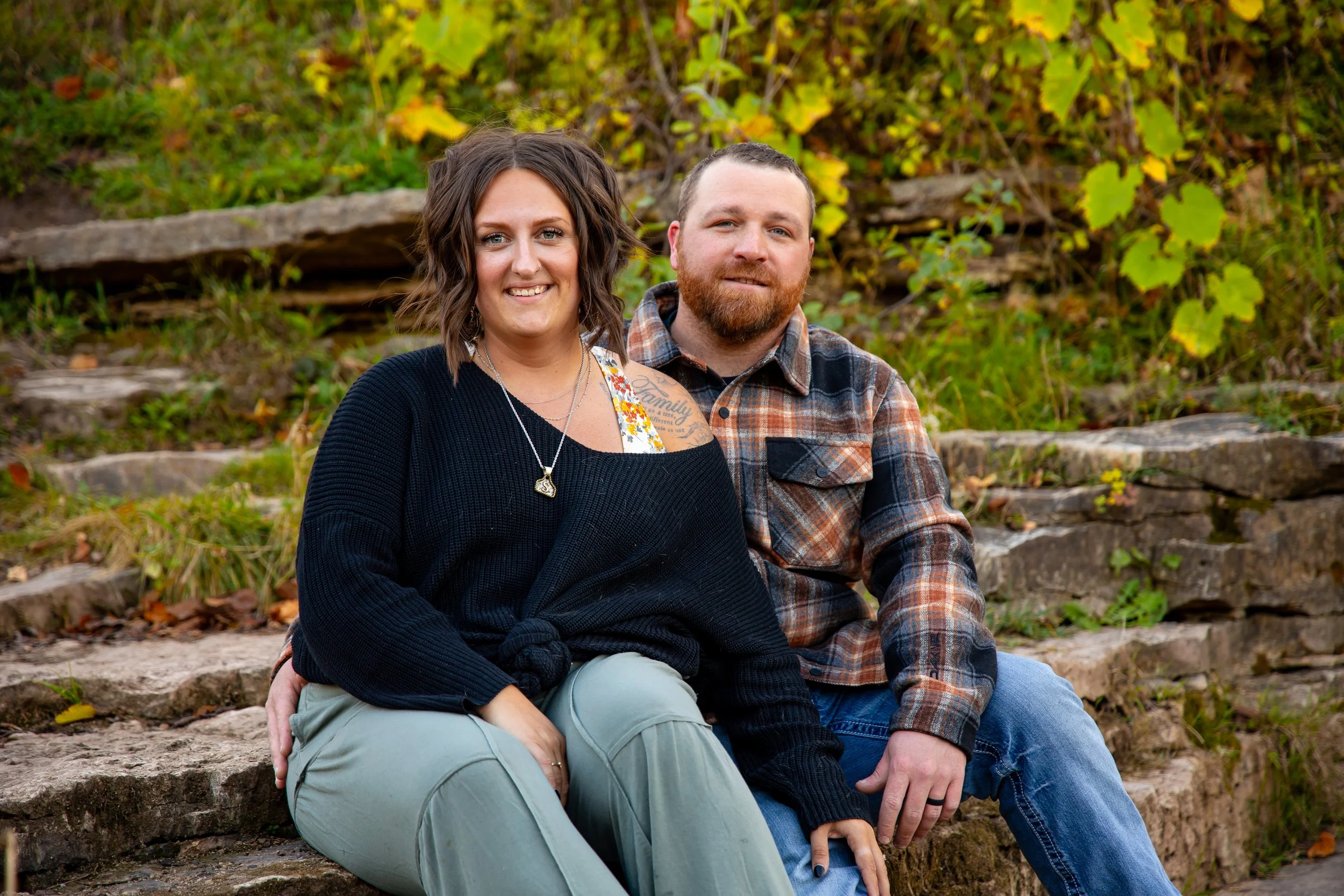 A smiling woman with brown hair and a man with a beard sitting outdoors on stone steps surrounded by green and yellow foliage.