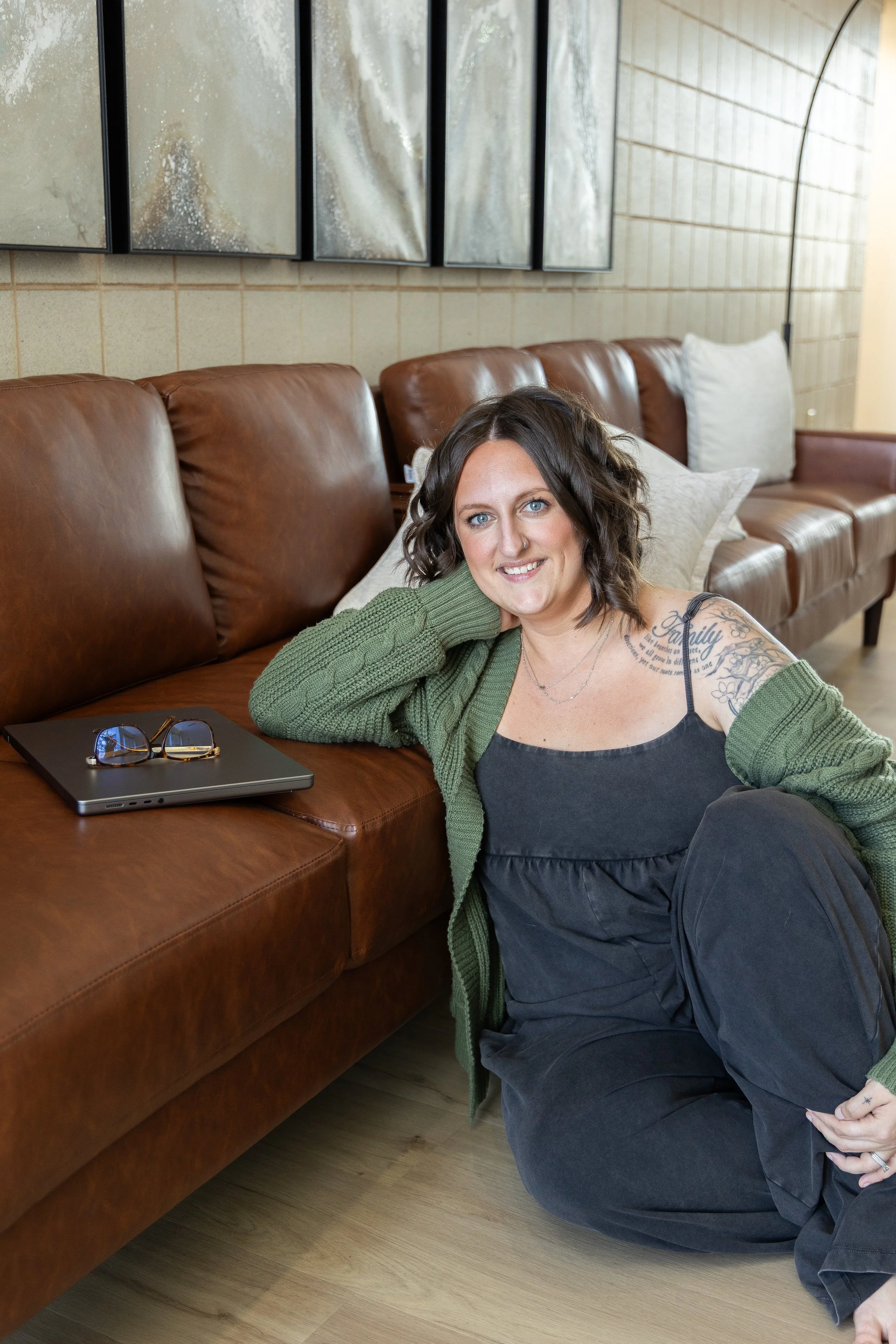 A woman with dark hair and tattoos sitting on the floor next to a brown leather couch, smiling at the camera in a room with beige tiled walls and a wooden floor.